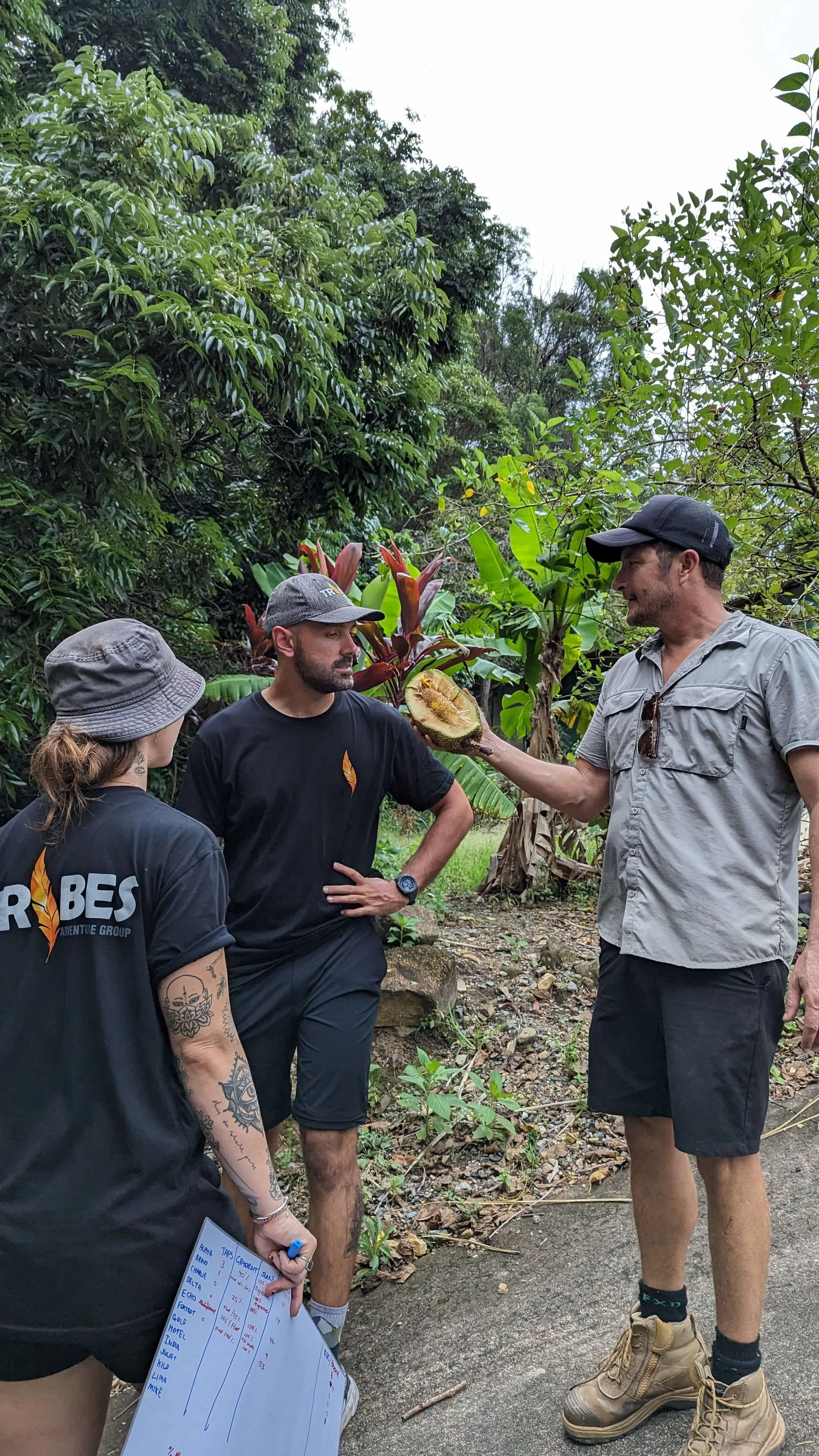 Three people in outdoor jungle setting, one man holding a large fruit, possibly a durian, while others observe. They are dressed in casual outdoor clothing, with one person holding a whiteboard or chart.