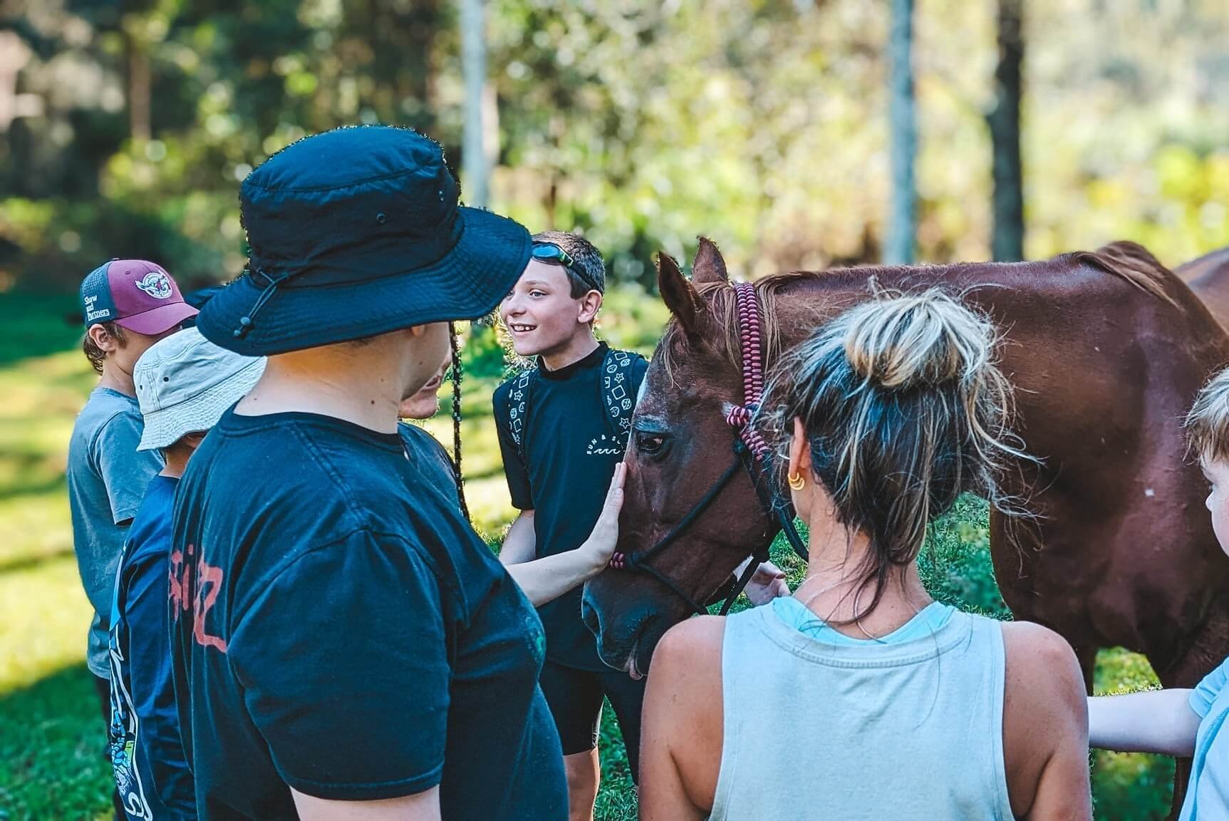 A group of children and adults outdoors petting a brown horse with white markings on its face in a wooded area.