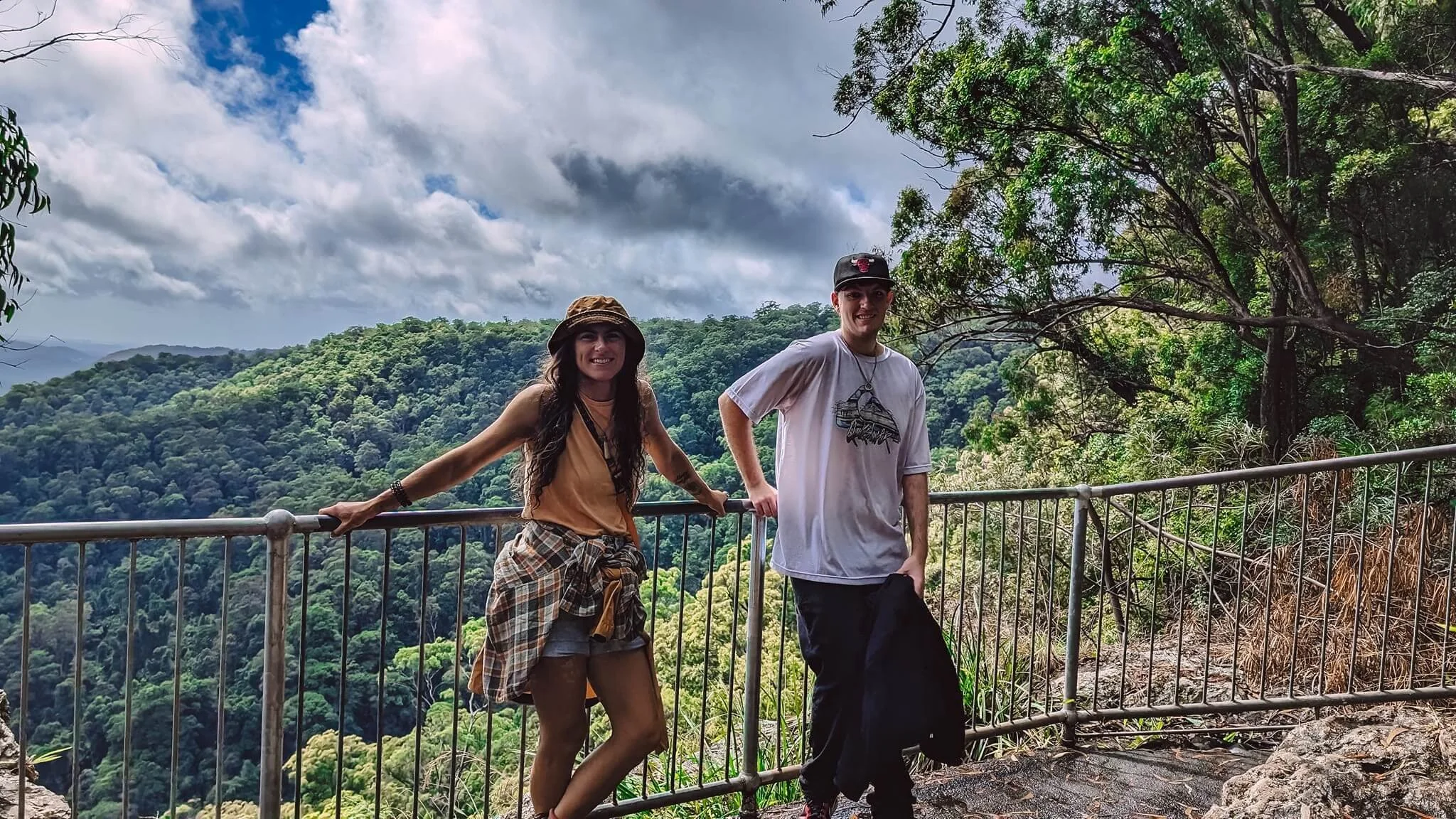 Two young adults, a woman and a man, standing on a viewing platform with a metal railing, overlooking a lush green mountain landscape under a cloudy sky. The woman has long dark hair, a tan bucket hat, and is dressed casually in shorts and a tank top. The man is wearing a black baseball cap, a white t-shirt with a graphic, and dark pants.