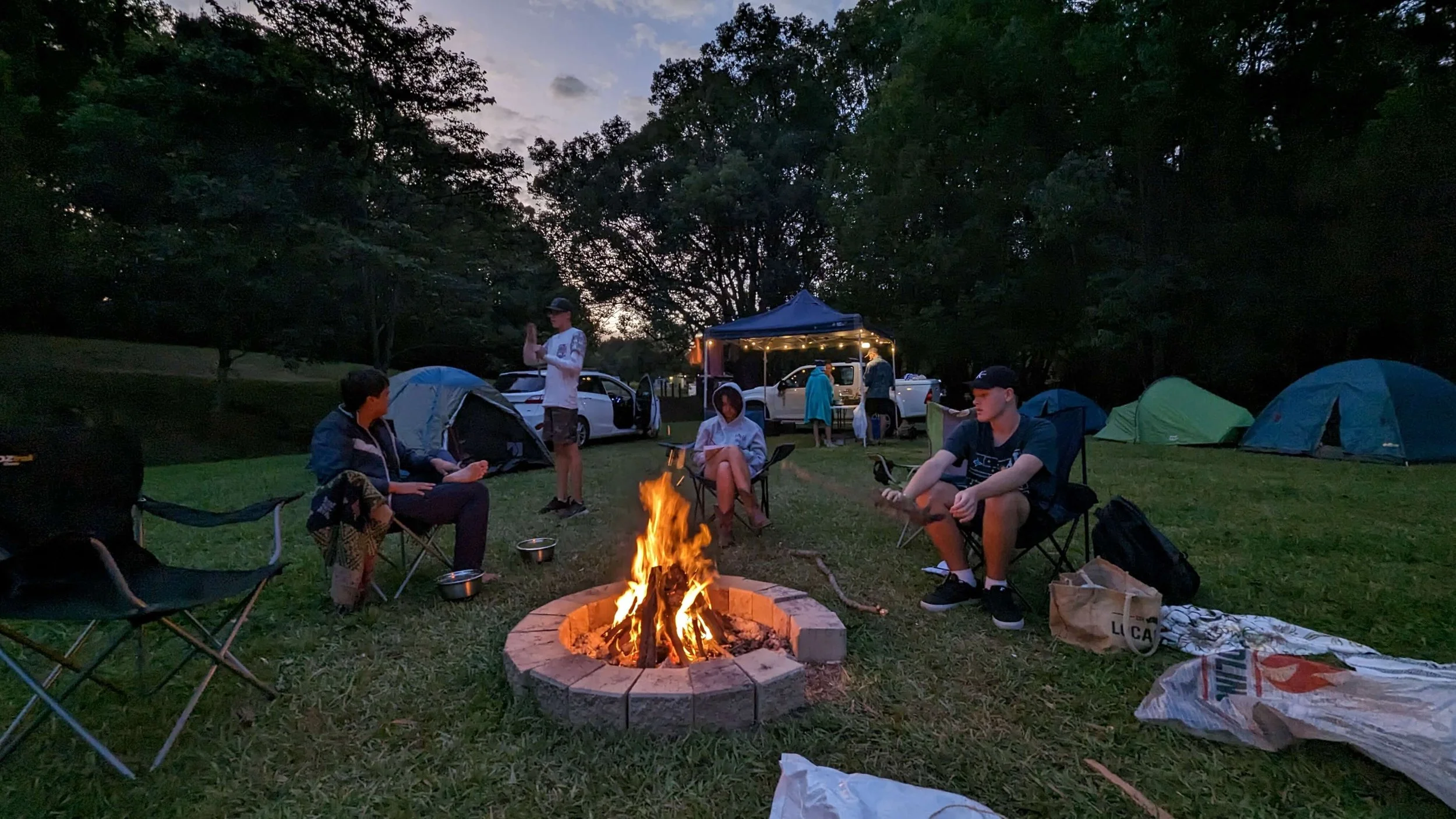 A group of people gathered around a campfire in a park during dusk, with tents, cars, and trees in the background.