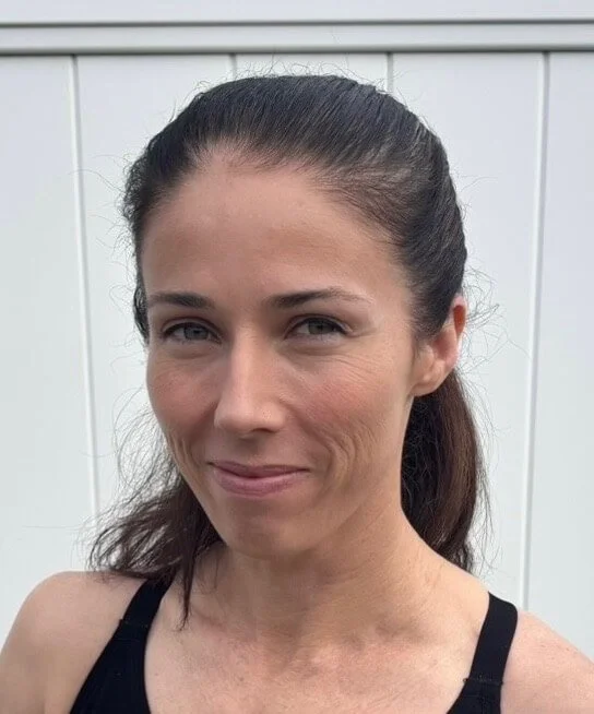 A woman with brown hair tied back, wearing a black tank top, standing in front of a white wooden fence.
