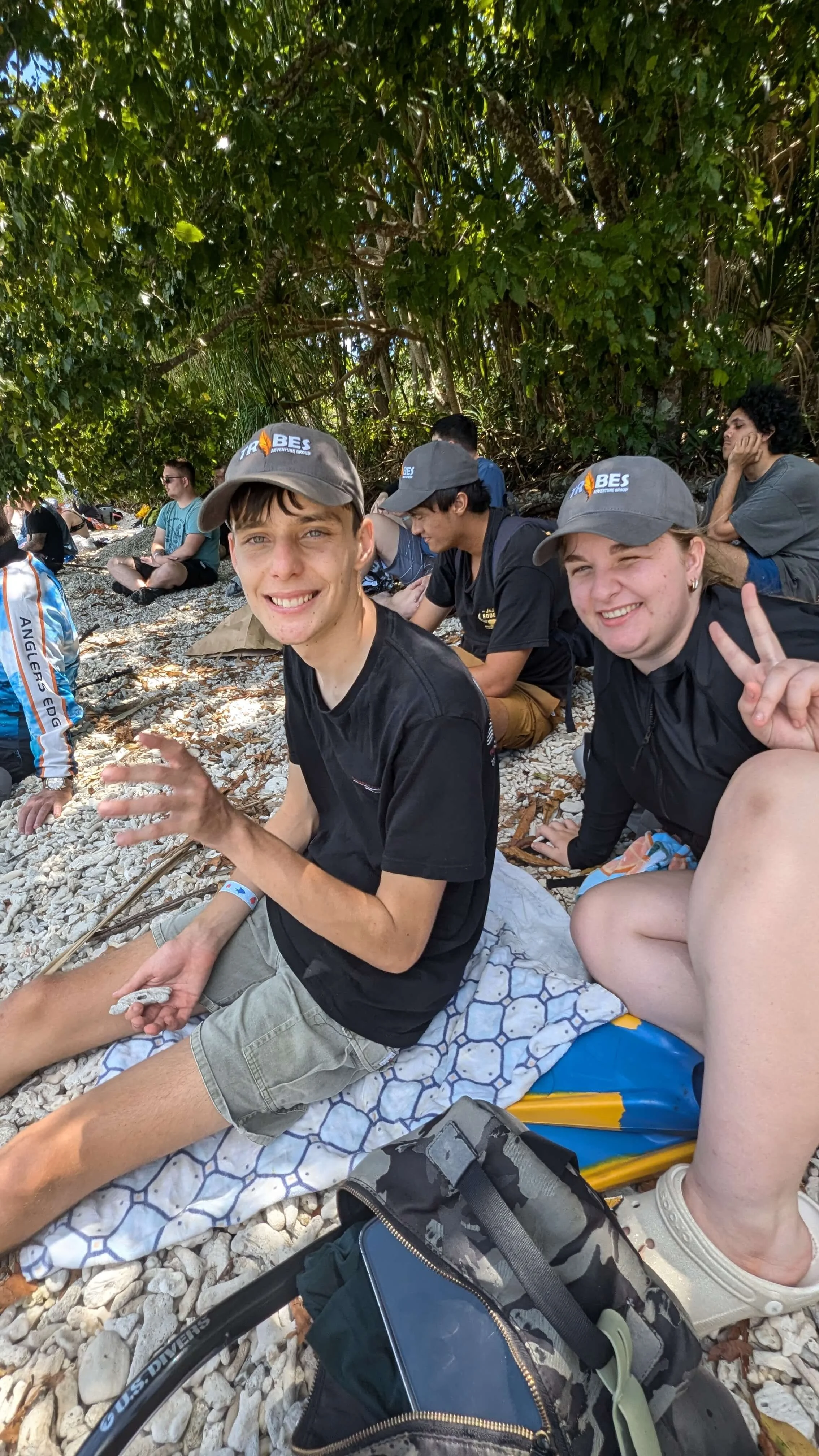 Group of young people sitting on a rocky beach under the shade of trees. Two people in the foreground are smiling at the camera; one giving a peace sign. They are wearing casual clothes and hats. There are backpacks and a hockey stick on the ground nearby.