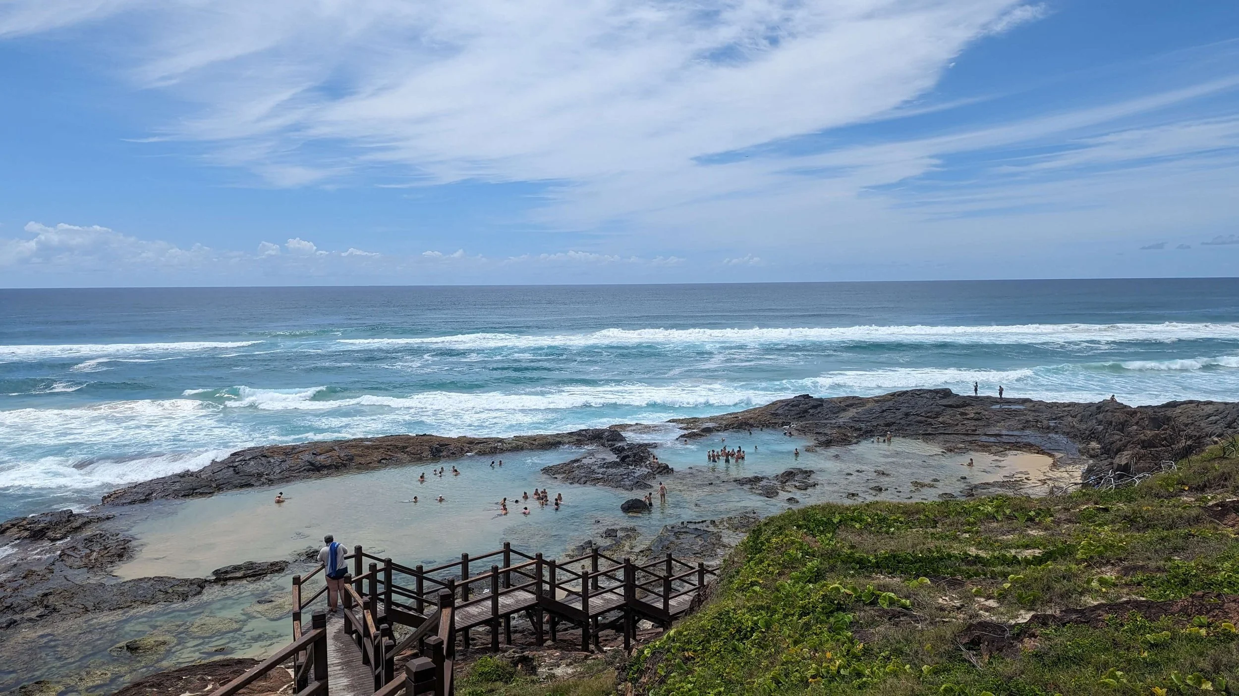 Beach with people swimming and relaxing on rocks, ocean waves, blue sky with clouds.