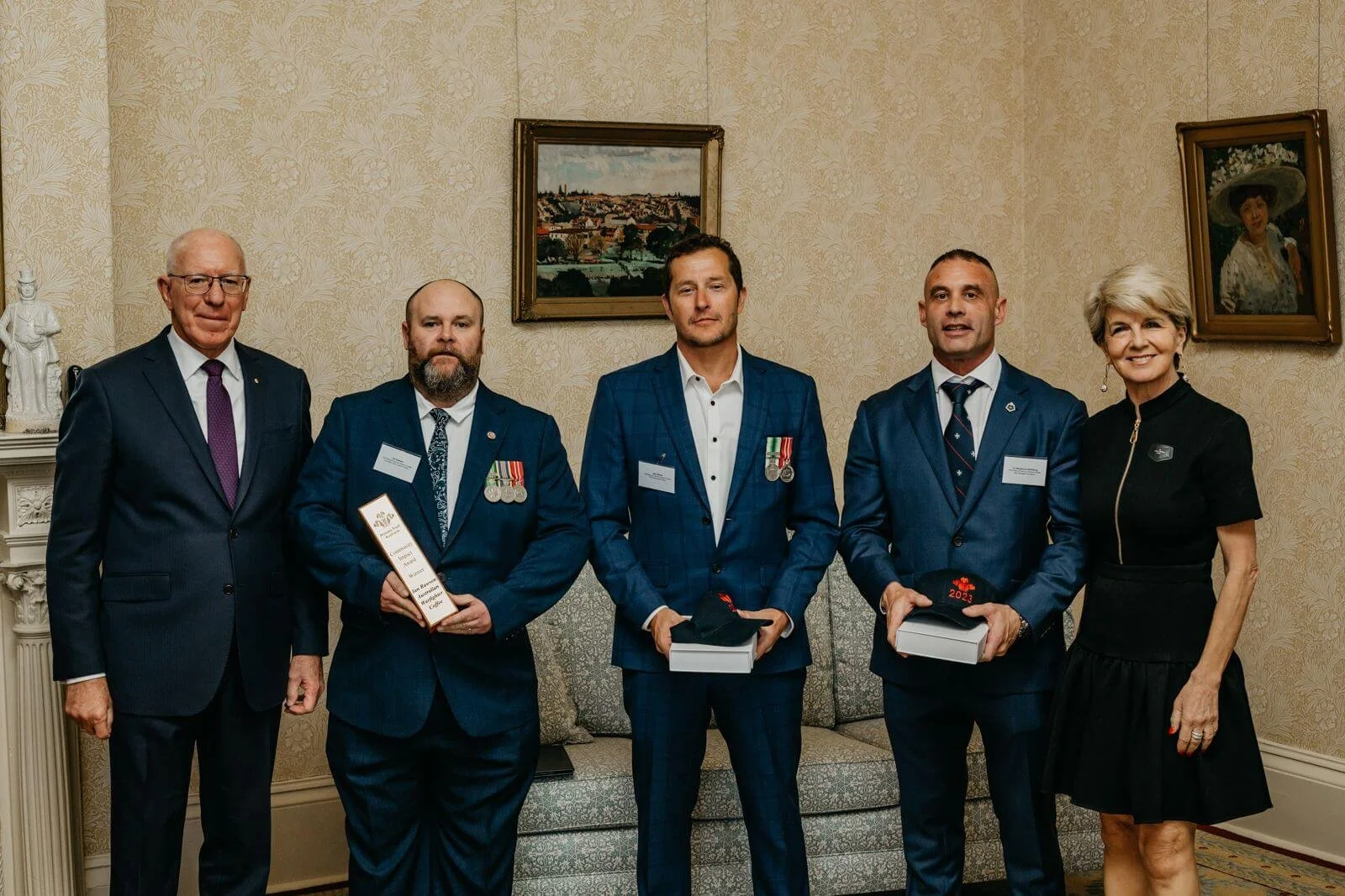 Five adults standing in a room, dressed in formal attire, with three men wearing medals and holding awards, and two women, one on each end, smiling at the camera. The room has wallpapered walls, framed artwork, and a small sofa.