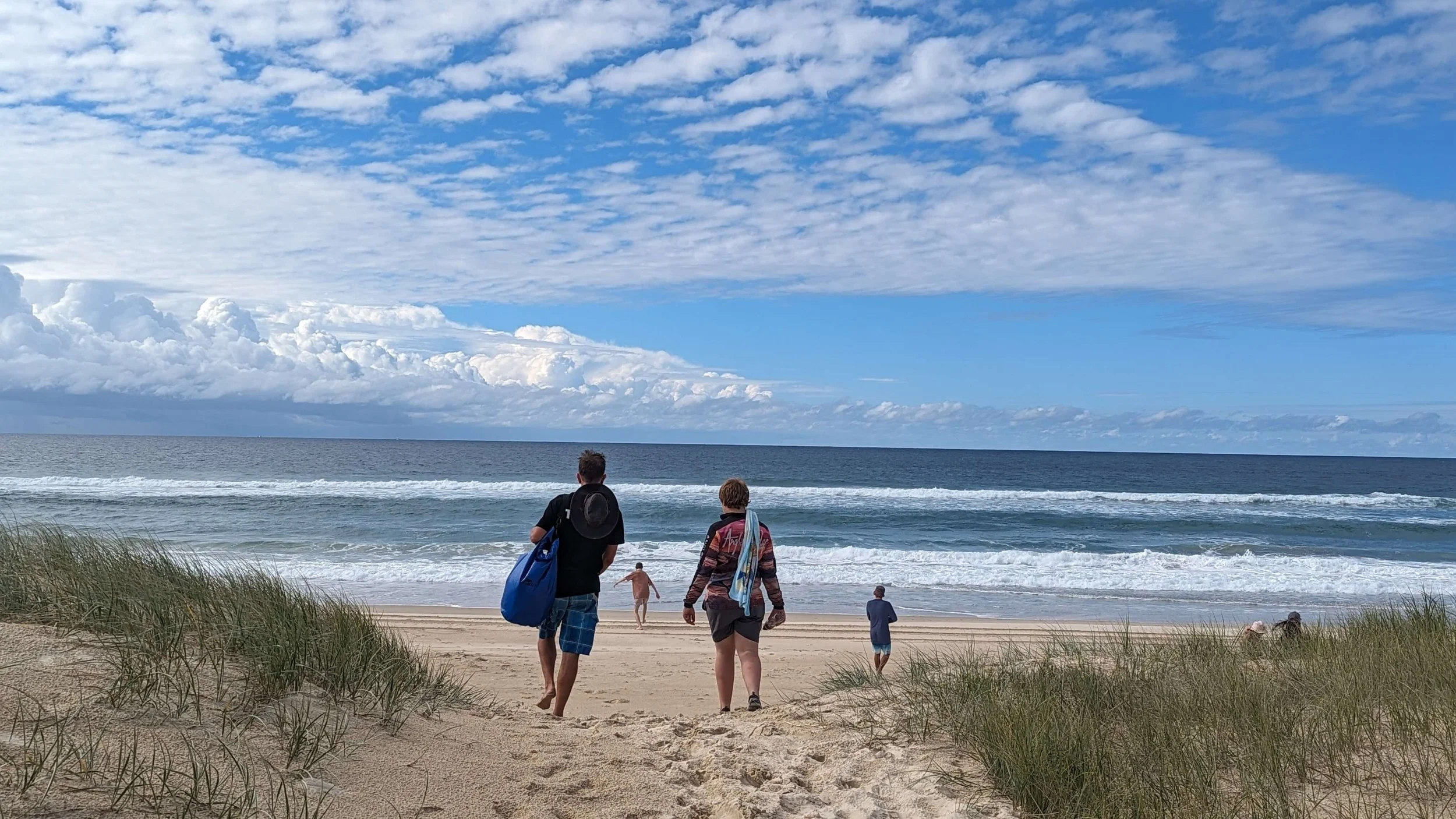 People walking on the sandy beach towards the ocean with blue sky and scattered clouds overhead.