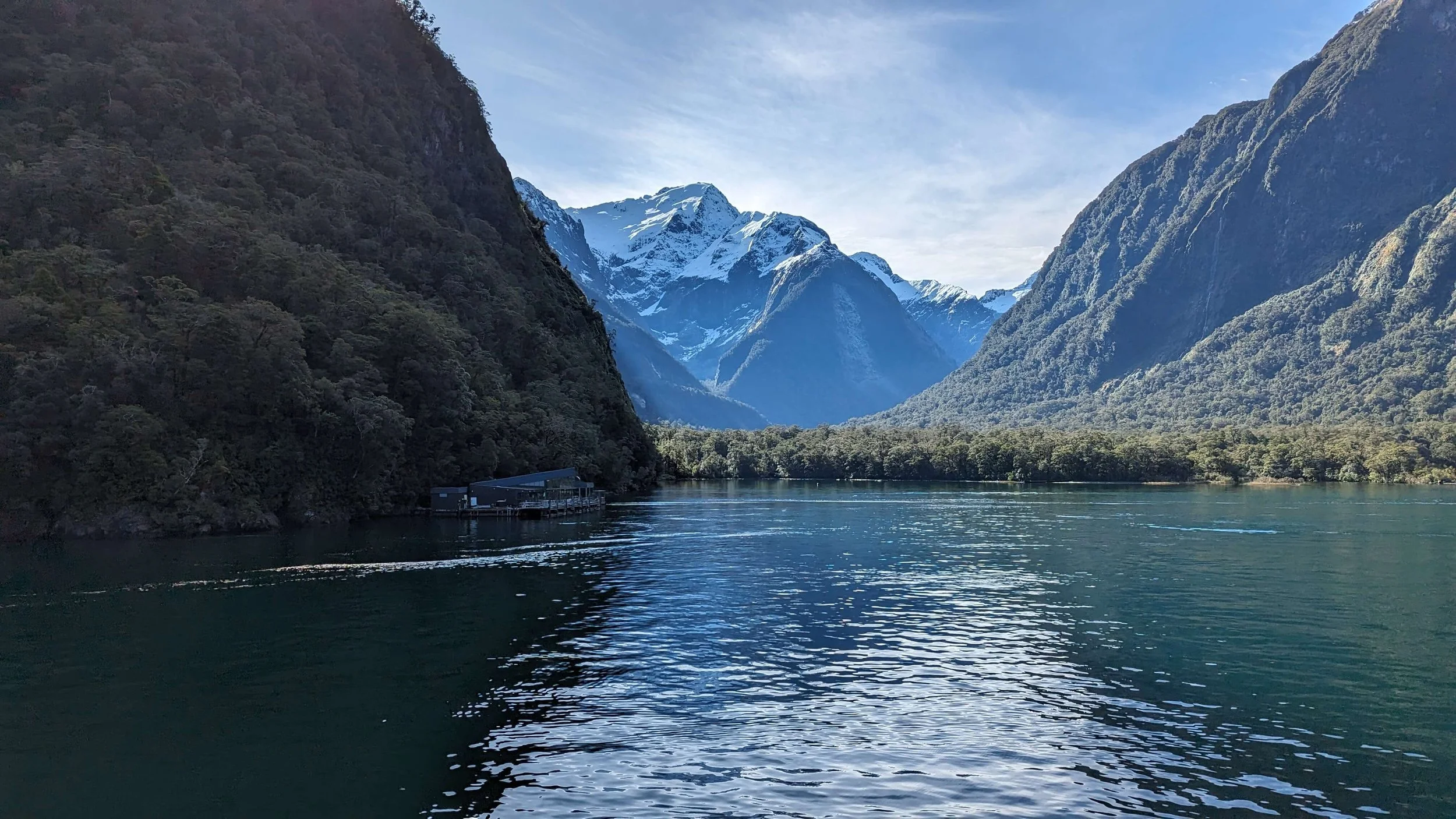 A scenic view of a lake surrounded by lush green mountains, some with snow-capped peaks, under a partly cloudy sky.