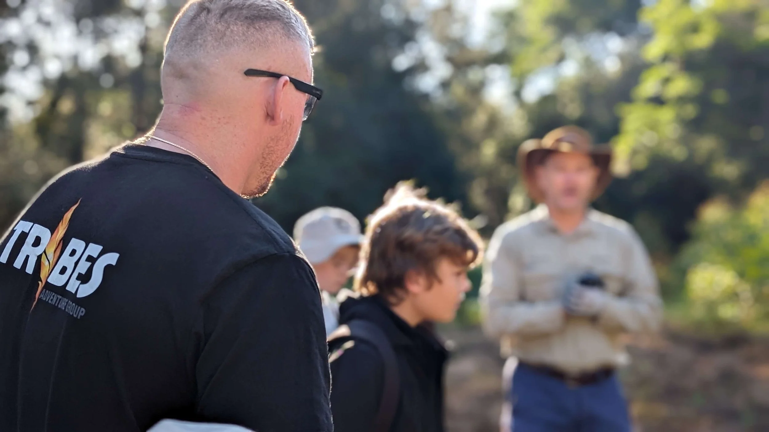 A group of people outdoors, with a focus on a man wearing black sunglasses and a black T-shirt, standing next to children and a woman in a wide-brimmed hat, with trees in the background.