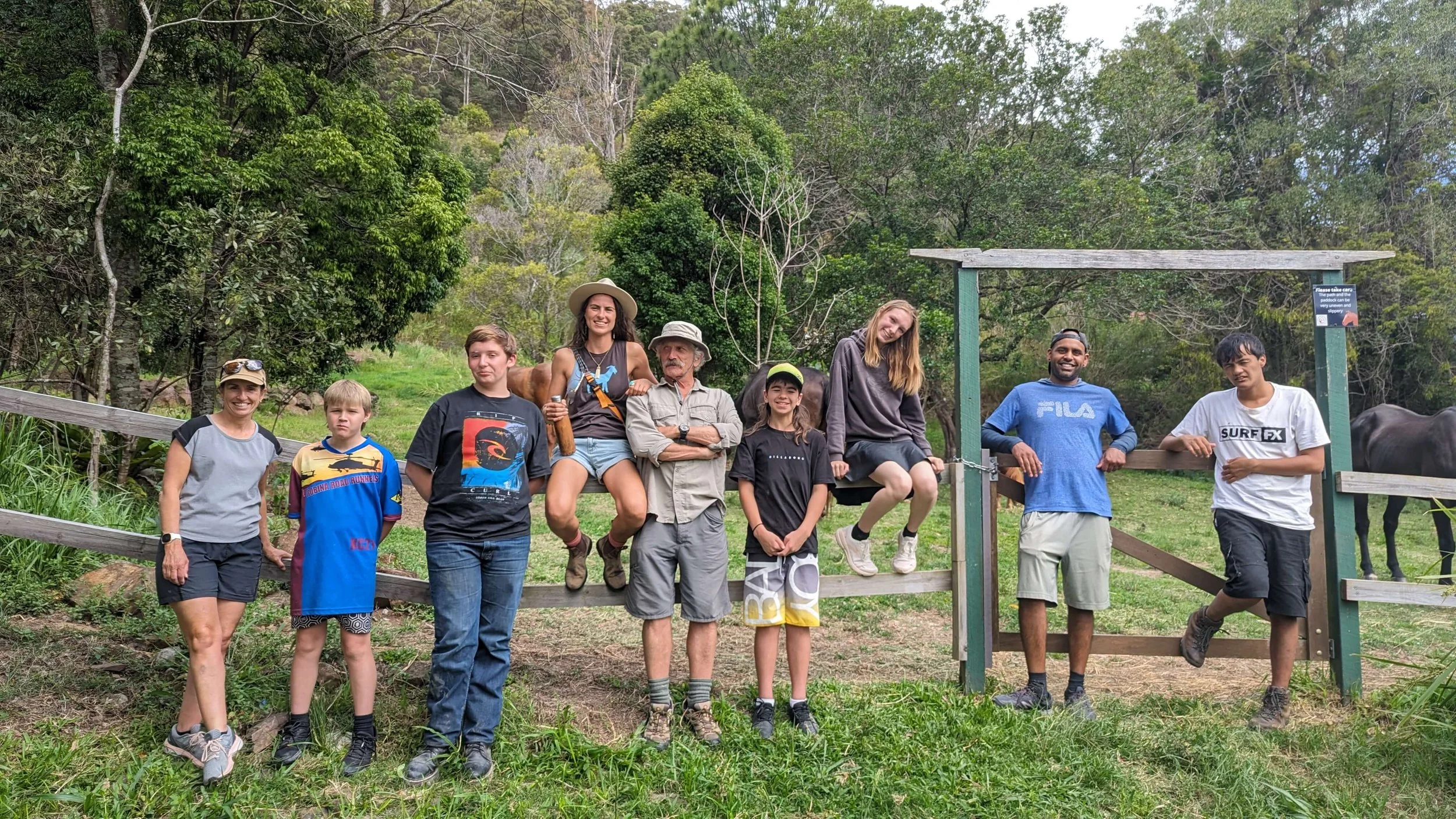 Group of people outdoors in a wooded area, standing and sitting on a wooden fence, with trees and greenery in the background.