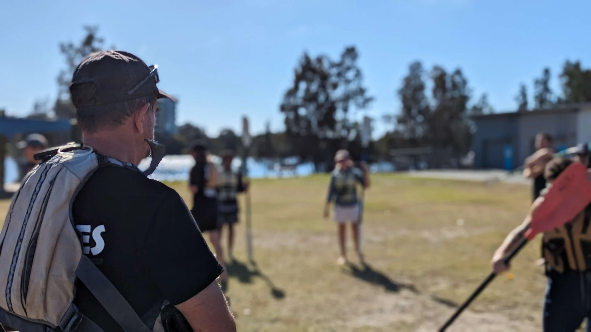 A man with a backpack looking at a group of people in an outdoor park with trees and blue sky.