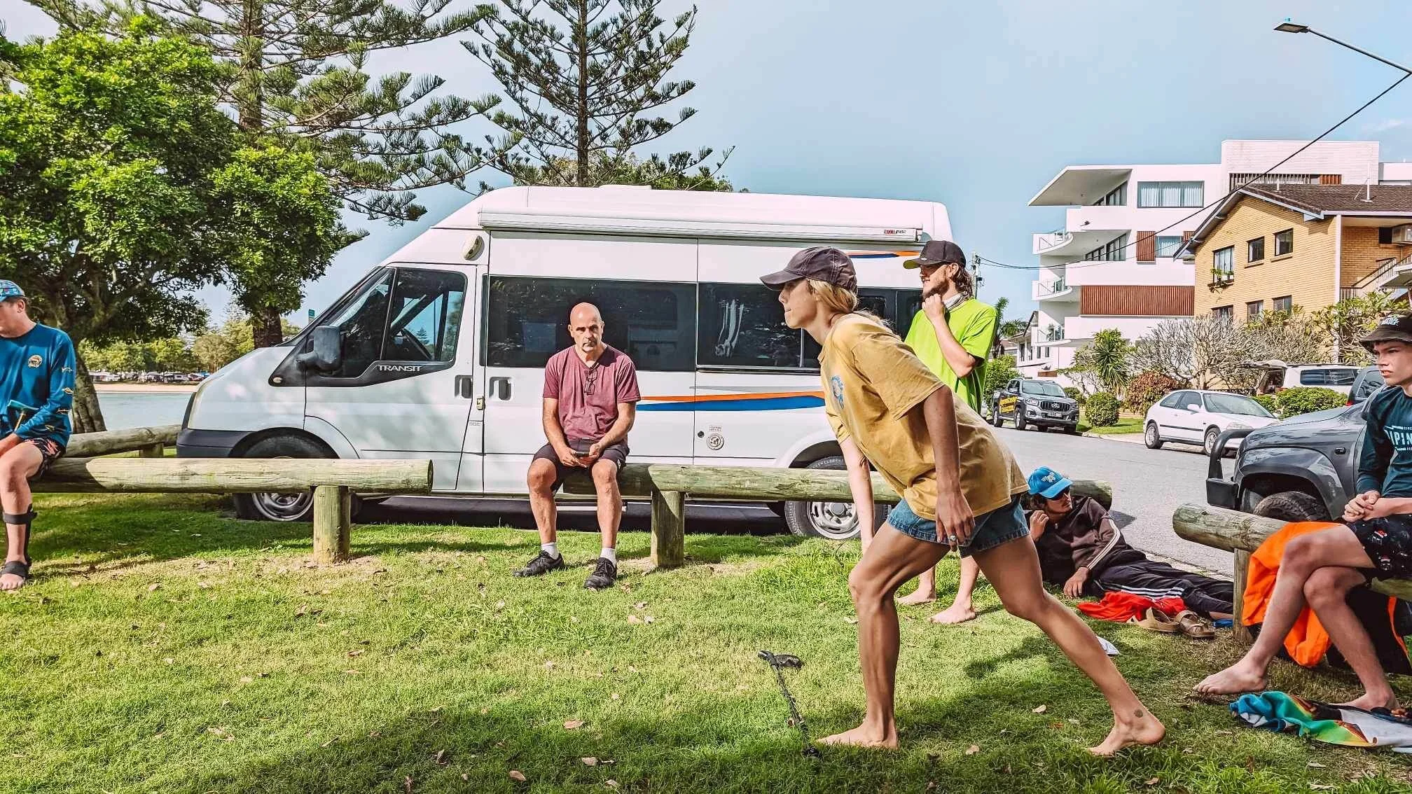 Several people, mostly young, gathered outdoors in a park-like area near a sidewalk and parked cars. Some are sitting, some standing, and one is walking. There is a white camper van in the background behind a wooden barrier, with trees and modern multi-story apartment buildings further behind.