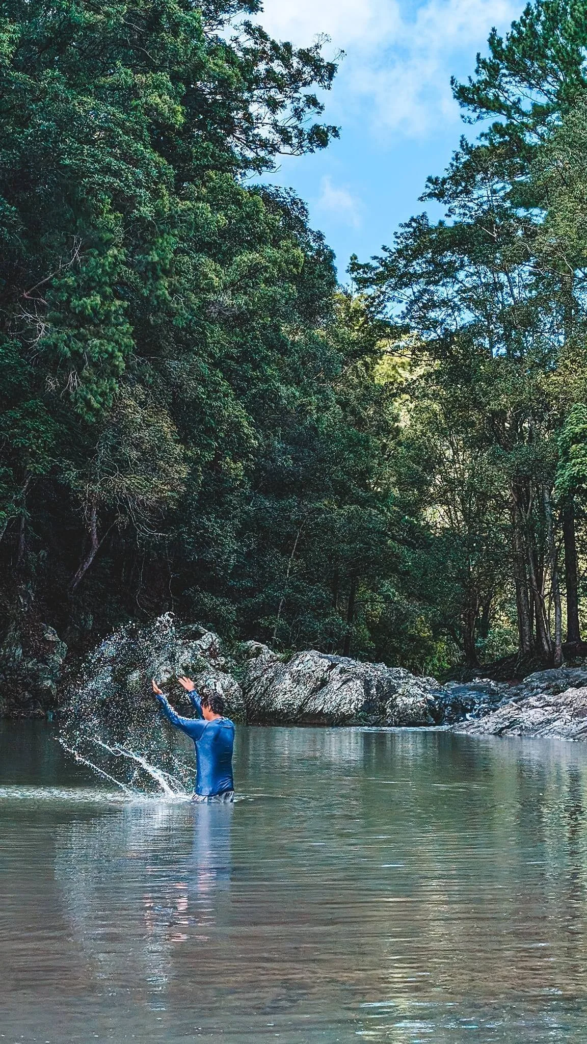 A person wearing a blue long-sleeve shirt and dark shorts is standing in a river, splashing water with their arms raised. The river is surrounded by dense green trees and rocks, with a partly cloudy sky overhead.