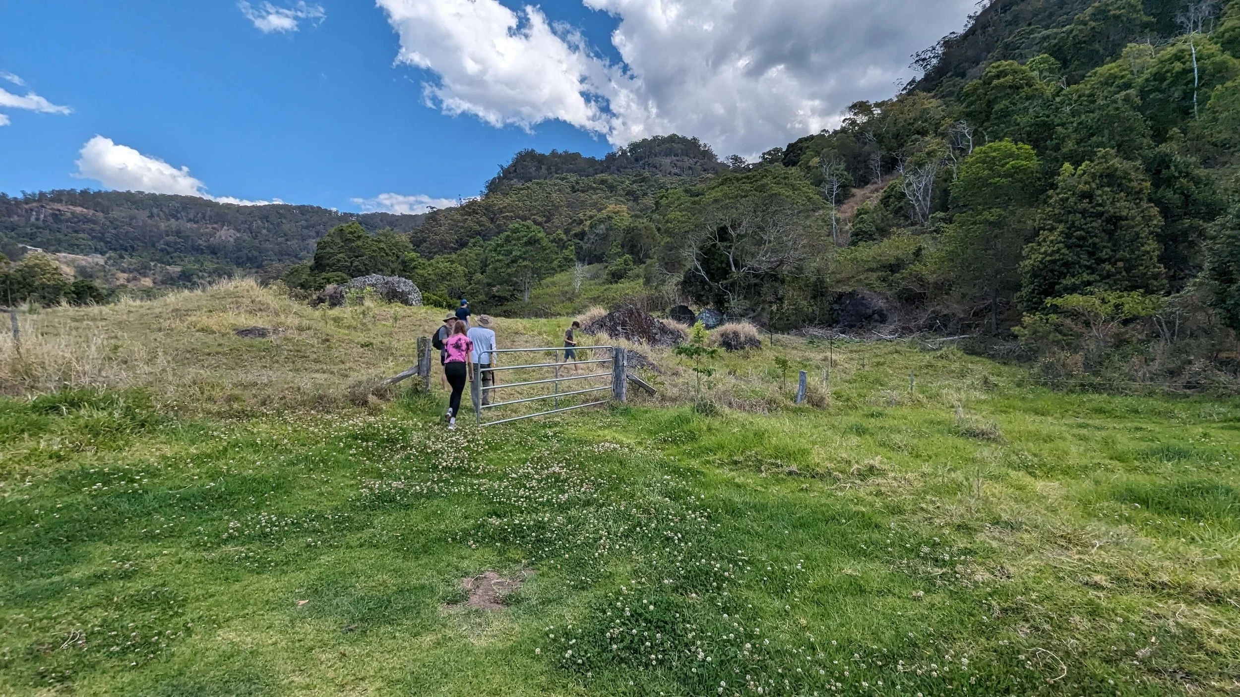 A group of people walking through a grassy field with a metal gate, surrounded by green hills and trees under a partly cloudy sky.