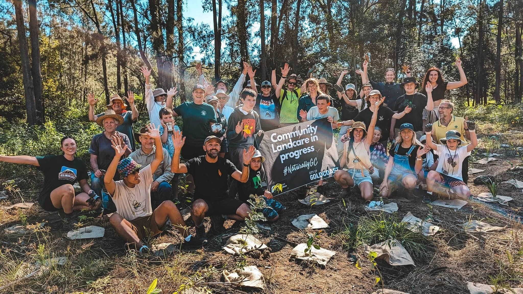 A large group of people from diverse backgrounds gathered in a forest for a community landcare event, holding a banner that reads 'Community Landcare in progress #NaturallyGC City of Gold Coast,' smiling and raising their hands.