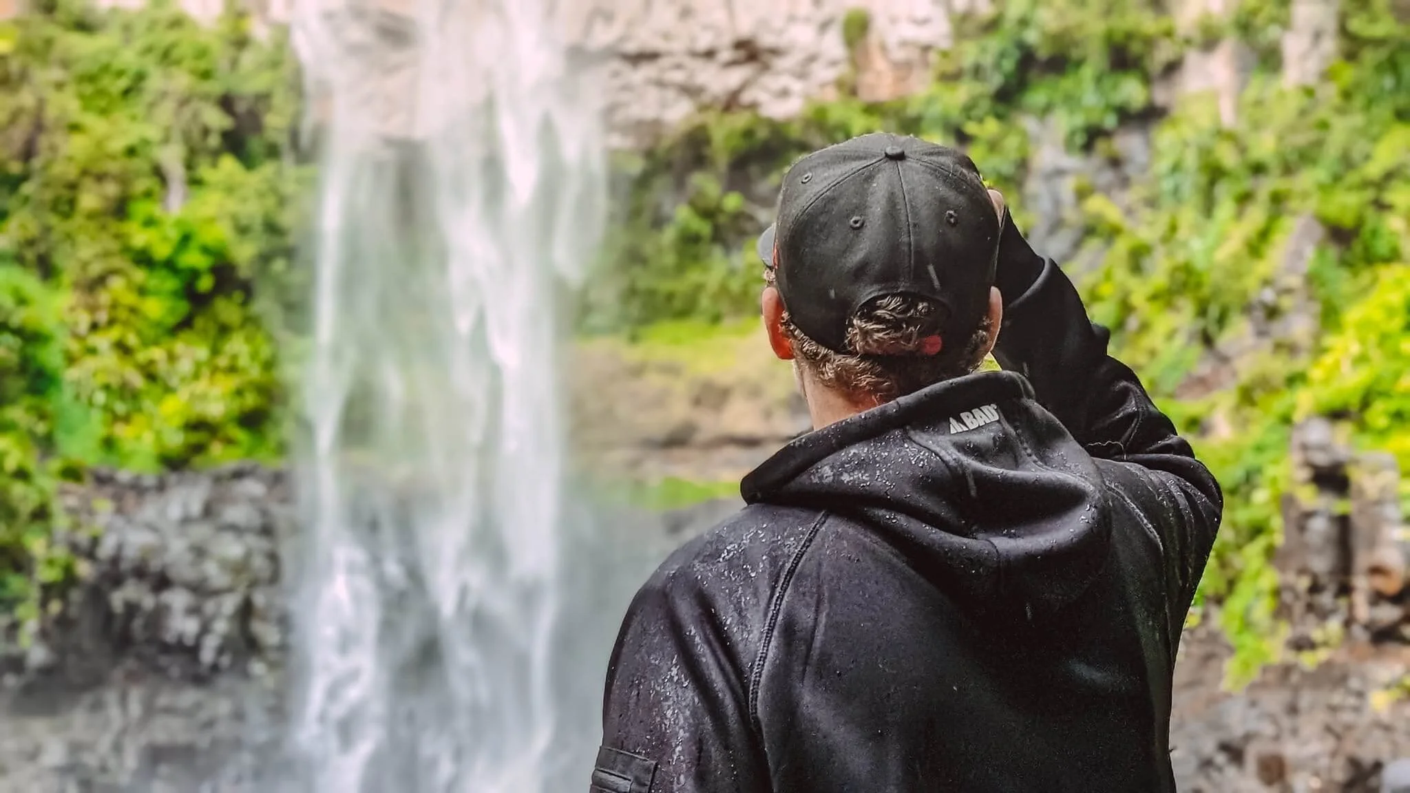Person in a black raincoat and black cap facing a waterfall in a lush green forest.