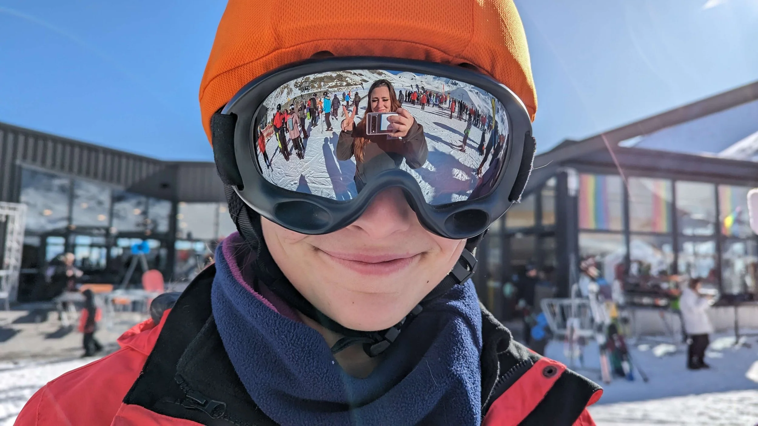 A person in ski gear and goggles taking a selfie that reflects a snowy outdoor ski resort with people in winter clothing in the background.