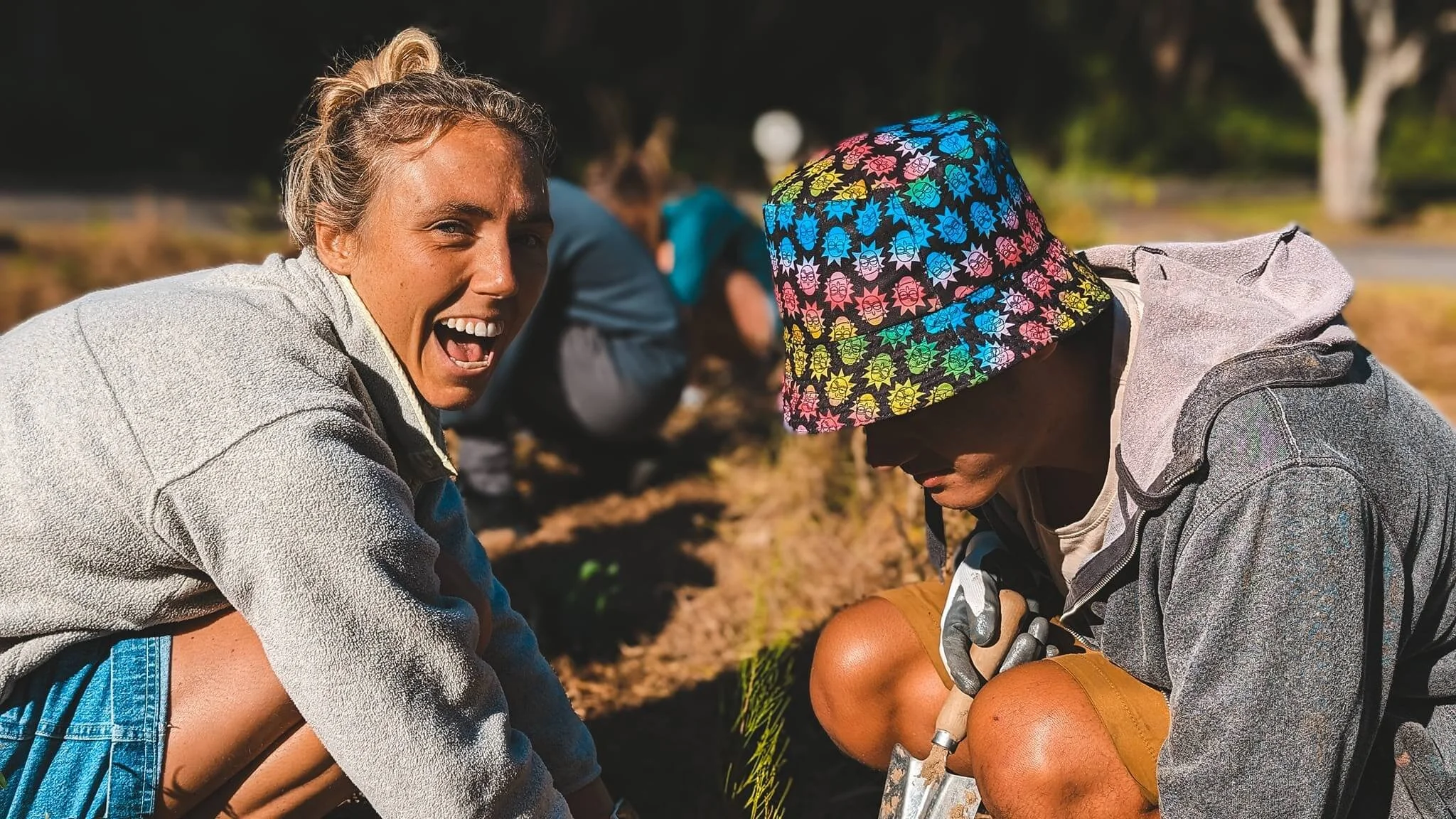 Two women outdoors working in a garden, one laughing and wearing a gray jacket, the other with a colorful sun hat and gray hoodie, focused on gardening.