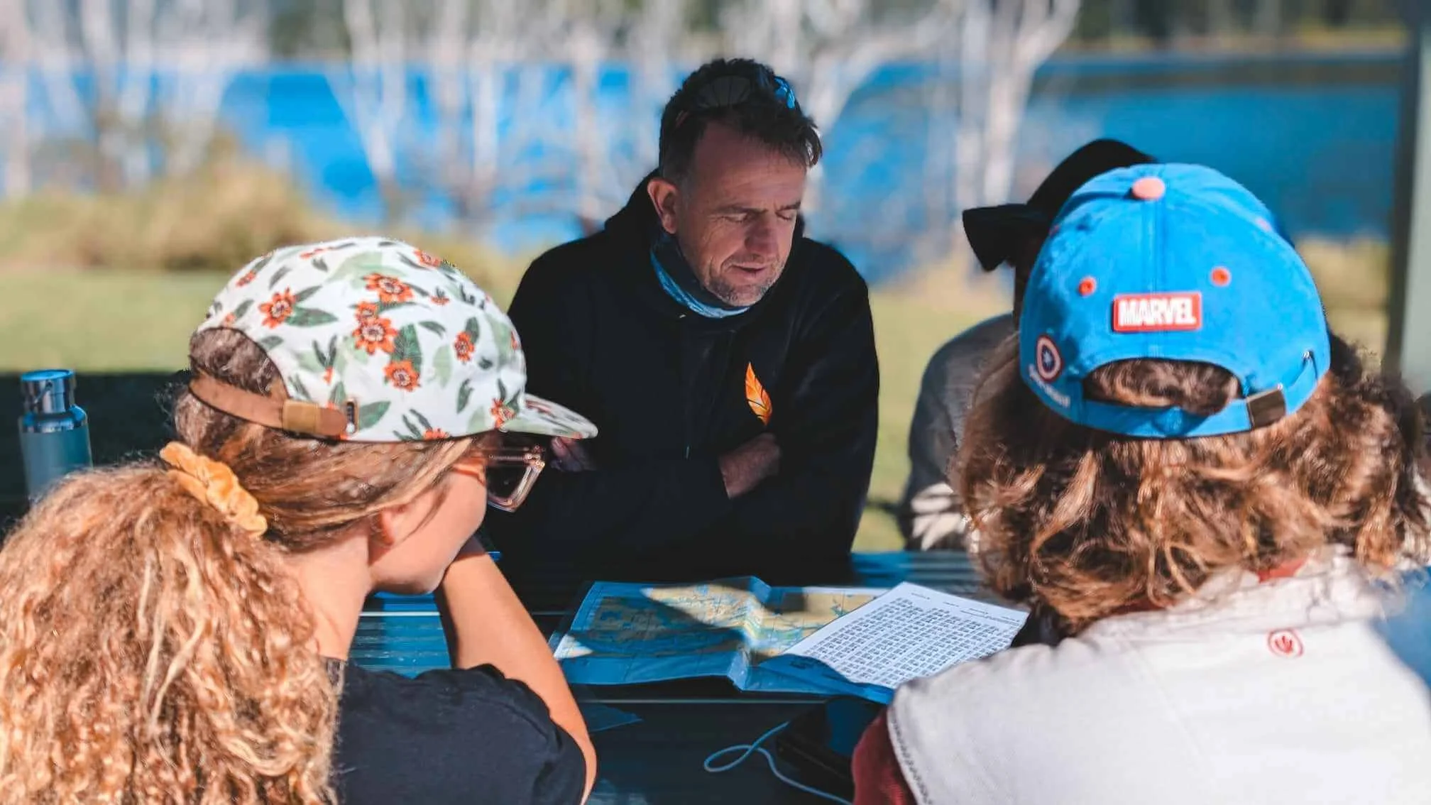 A man with dark hair and a beard, wearing a black jacket, is sitting at a table with three children. One girl has curly red hair, wearing a floral cap, and glasses, with her head down. Another girl with curly brown hair is wearing a blue Marvel cap. The group appears to be outdoors during daytime, with a blurred background of trees and blue structures, looking at a map on the table.