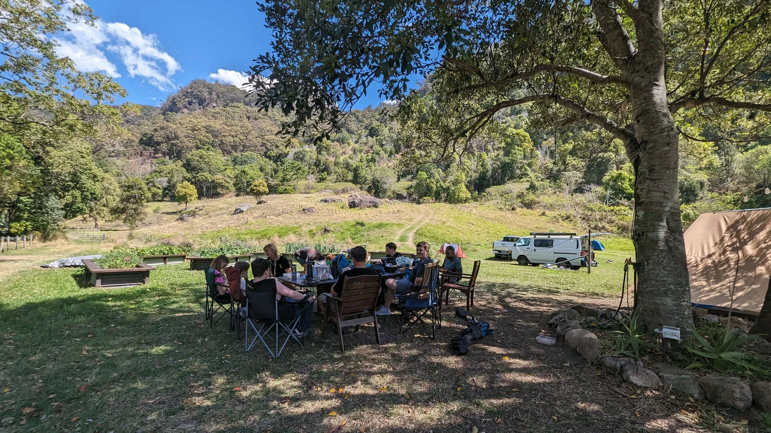 Group of people sitting around a picnic table under a large tree, with camping equipment and vehicles nearby in a scenic outdoor setting with hills and trees.