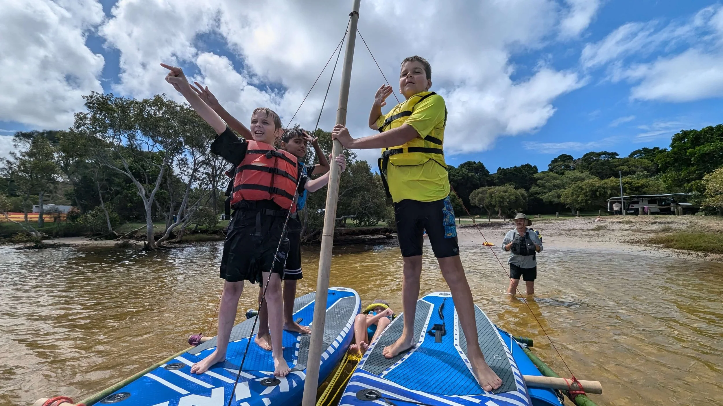 Children and an adult on a stand-up paddleboard in a river, holding a pole, with trees and a cloudy sky in the background.
