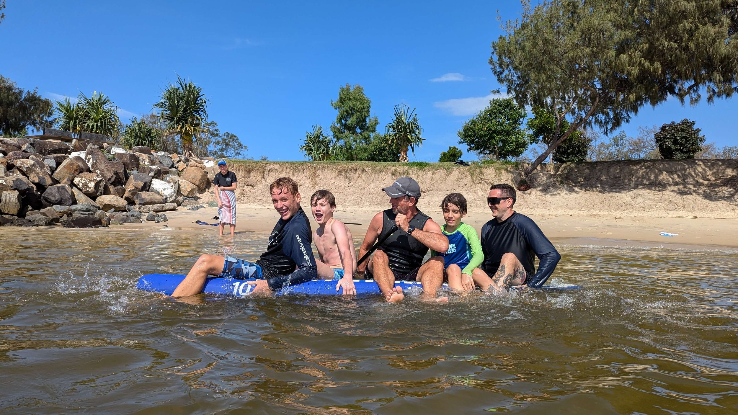 Group of six people sitting on an inflatable raft in a body of water, smiling and enjoying a sunny day at the beach with trees and a rocky shoreline in the background.