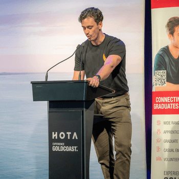 A man standing at a podium with the HOTA logo, speaking into a microphone at an event, with a large screen or banner in the background.
