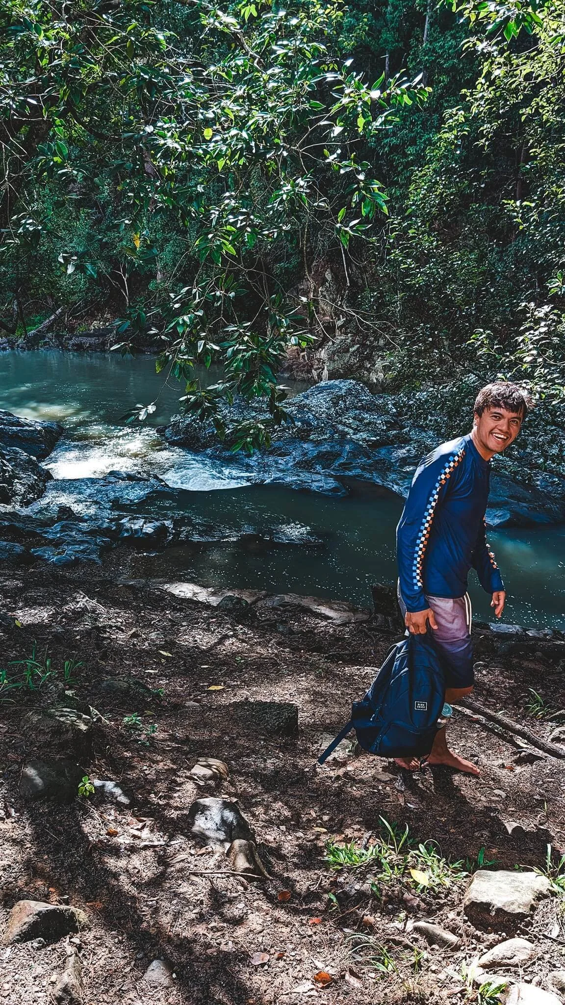 A young man in a blue long-sleeve shirt and gray shorts is standing on a muddy riverbank surrounded by lush green forest. He is smiling, holding a jacket in one hand, with a backpack on the ground.