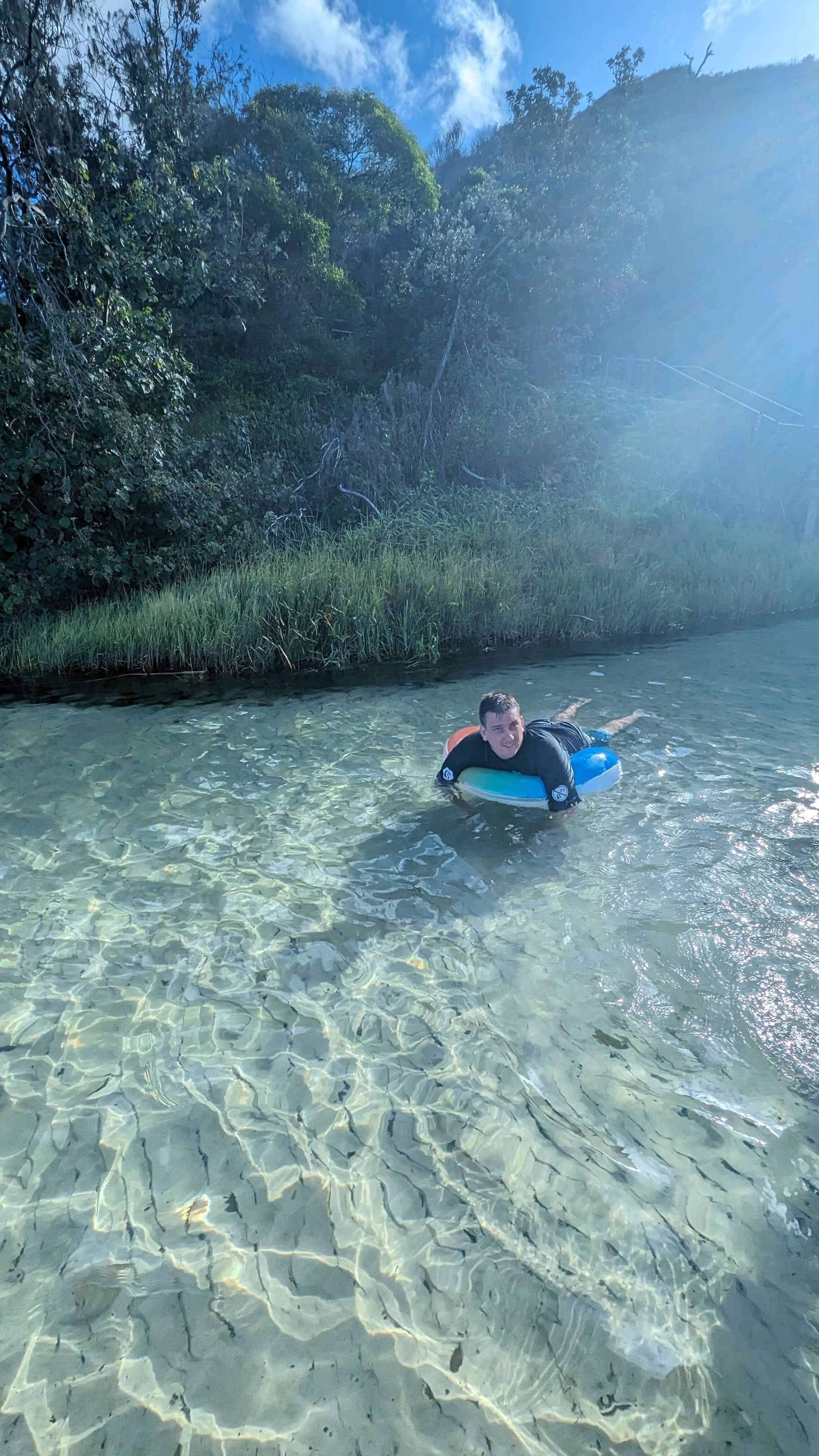 A person smiling and lying on a surfboard in clear shallow water near a grassy shoreline with dense trees and blue sky in the background.