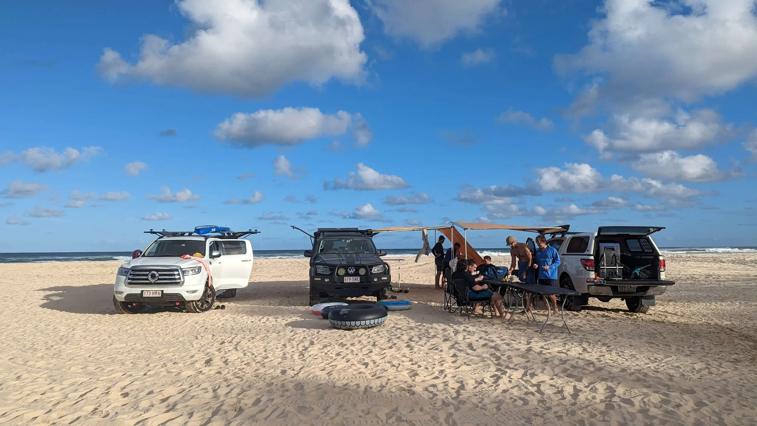 A group of people at the beach with three parked vehicles, some seated and others standing around a table, with a clear blue sky and clouds overhead.