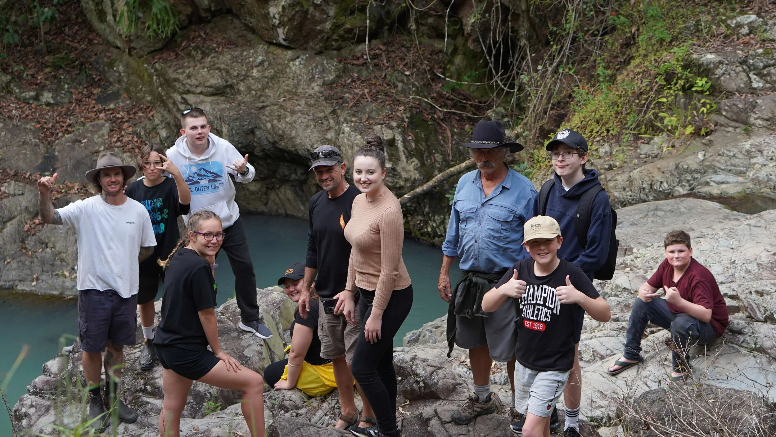 Group of people hiking in a rocky outdoor area near water, surrounded by trees and rocks.