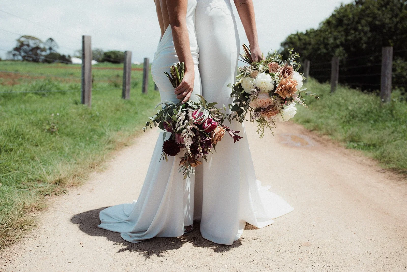 Two women in white dresses standing on a dirt path, holding bouquets of flowers, with a rural fence and green trees in the background.