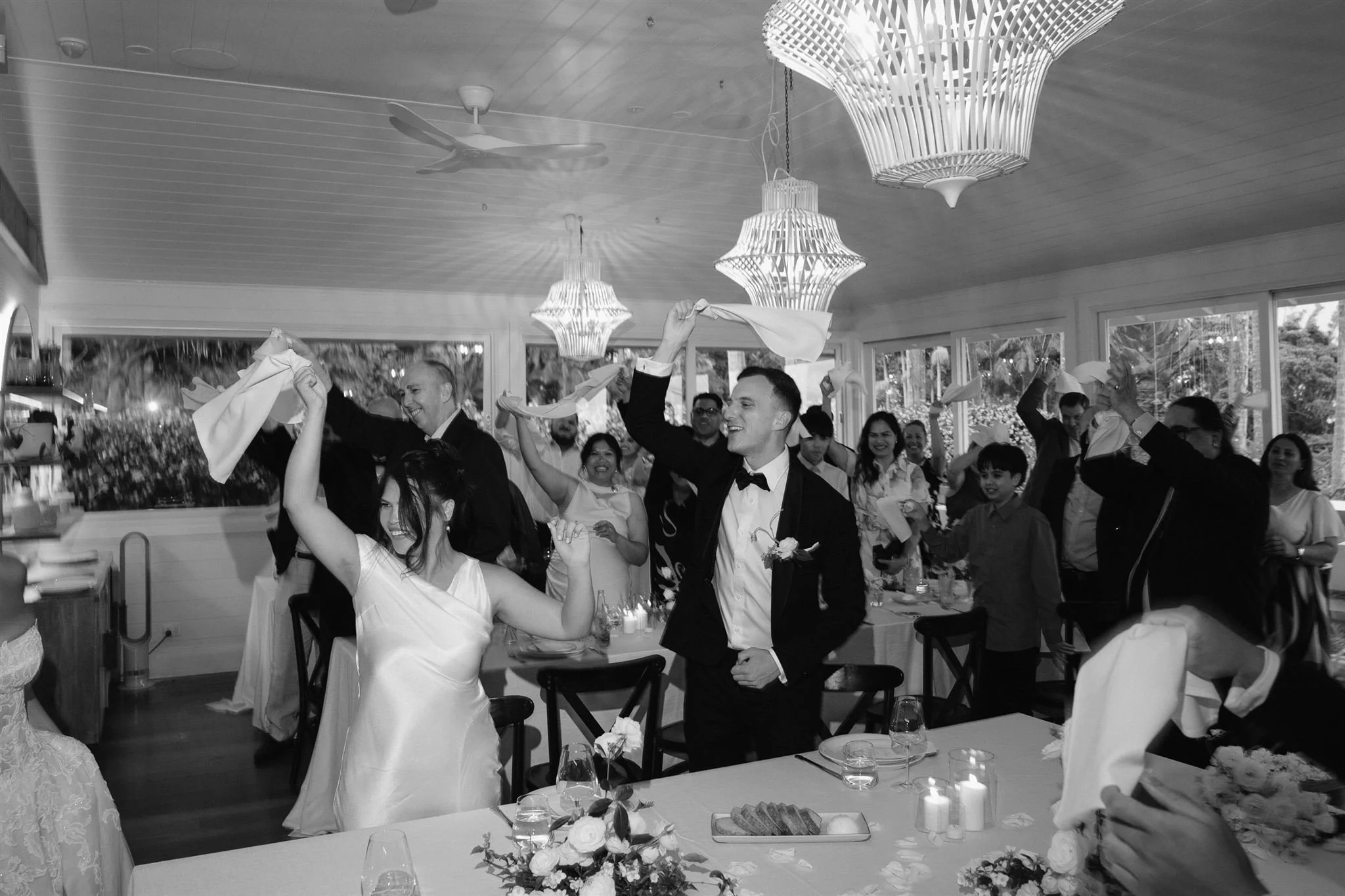 People dancing and celebrating at a wedding reception indoors, with window views of trees outside, decorated with hanging lamps and floral centerpieces on tables.