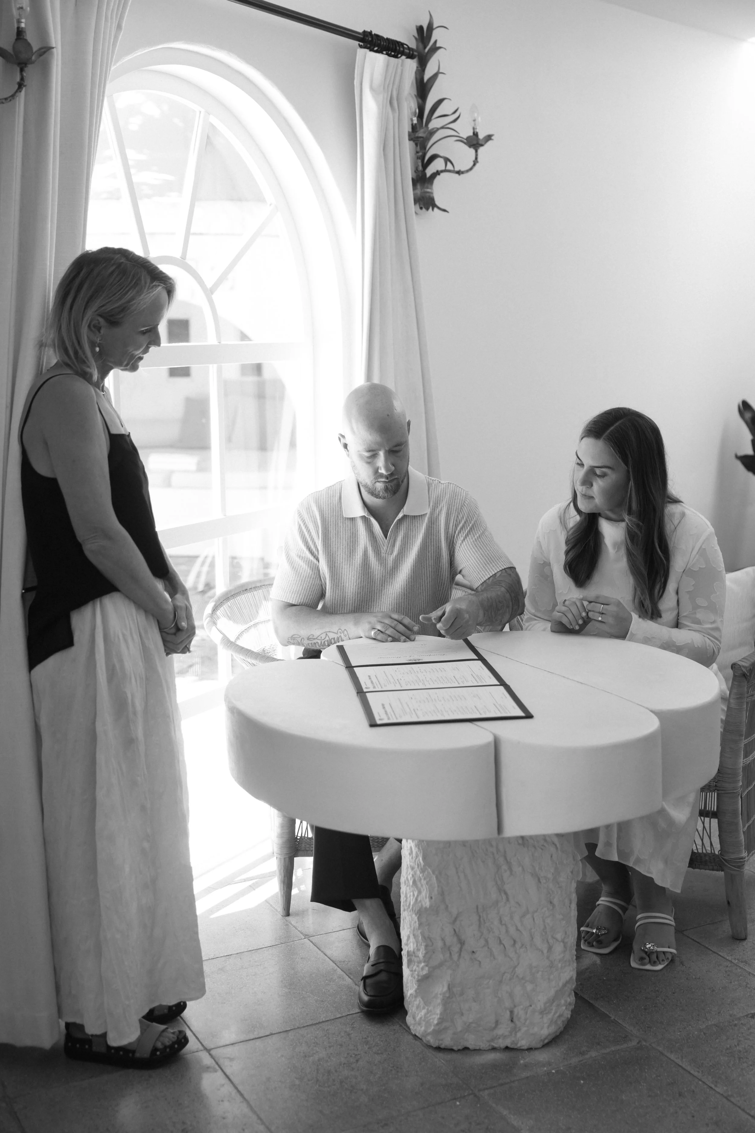 Three people sitting around a round table with a large rock base, looking at a menu or document. One woman standing, wearing a sleeveless top and a long skirt, two seated, one man with a shaved head and a beard, the other a woman with long hair, all indoors near a window with curtains.