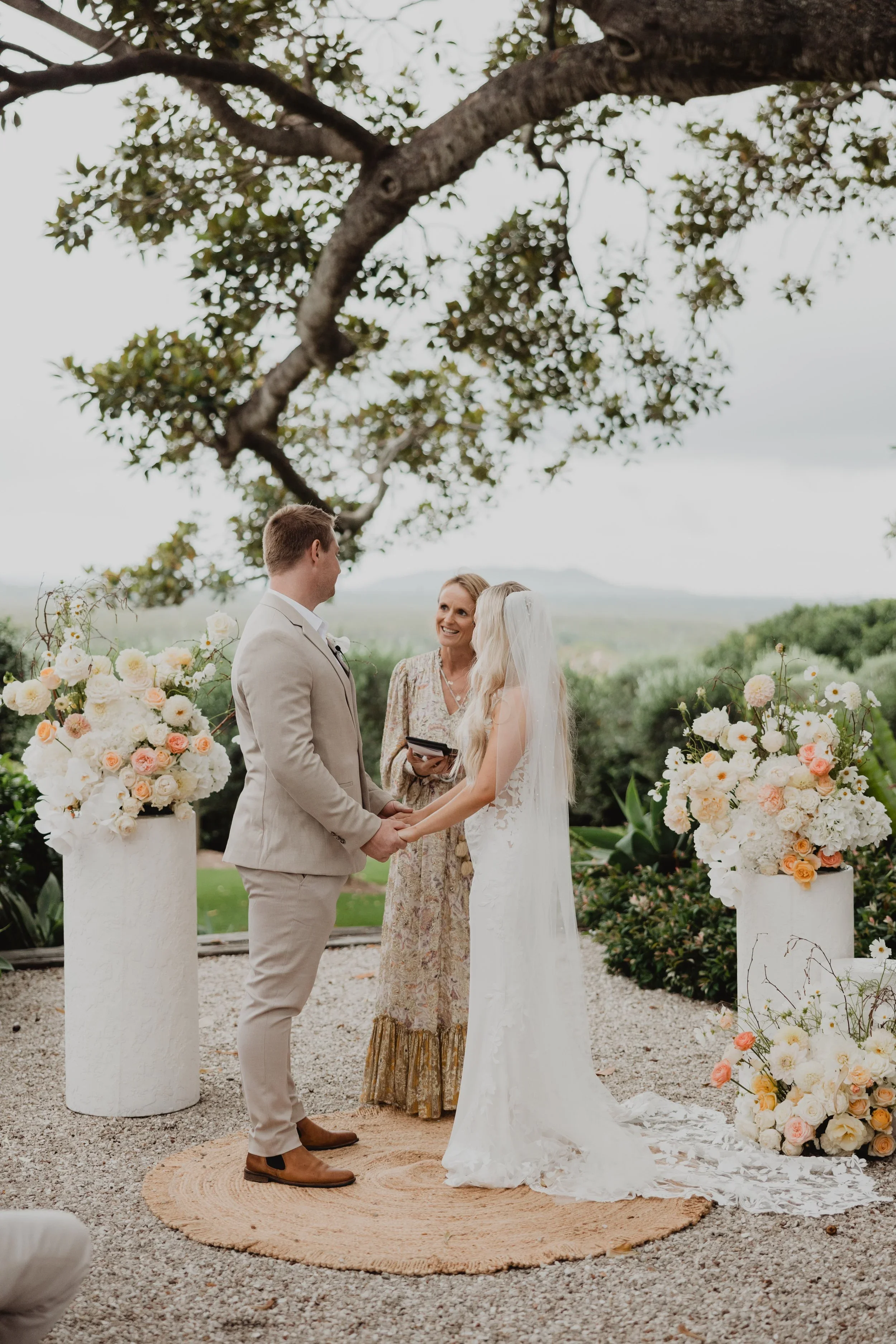A couple getting married outdoors under a large tree, with a woman officiating the ceremony. The bride is wearing a white wedding dress with a veil, and the groom is dressed in a beige suit. They are holding hands in front of floral arrangements in white planters, with a scenic landscape in the background.