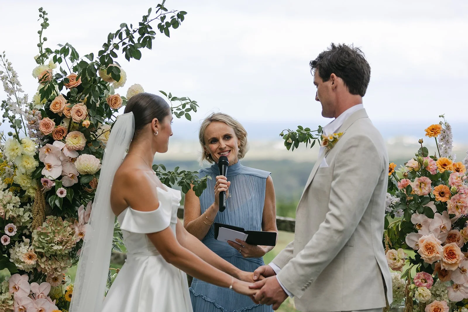 A wedding ceremony taking place outdoors with a bride and groom holding hands, facing each other, while a woman officiates and speaks into a microphone. Flowers and greenery surround them.