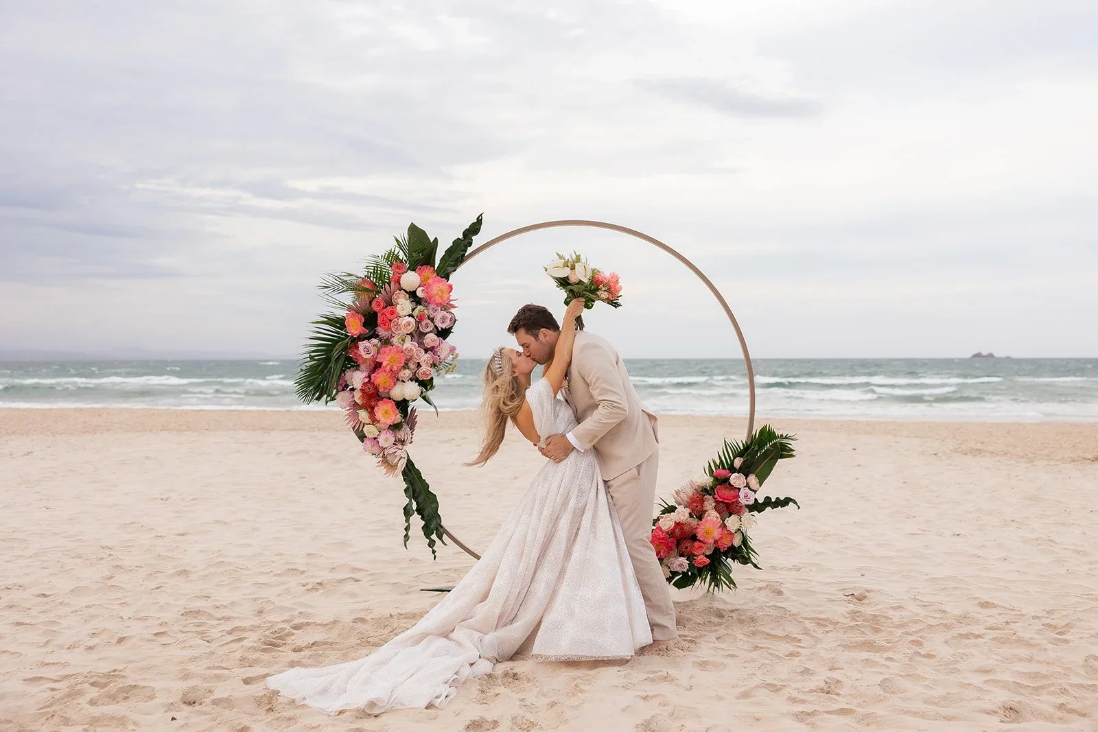 A bride and groom are kissing on the beach during their wedding ceremony, standing beneath a circular floral arch decorated with pink, white, and green flowers, with the ocean and cloudy sky in the background.