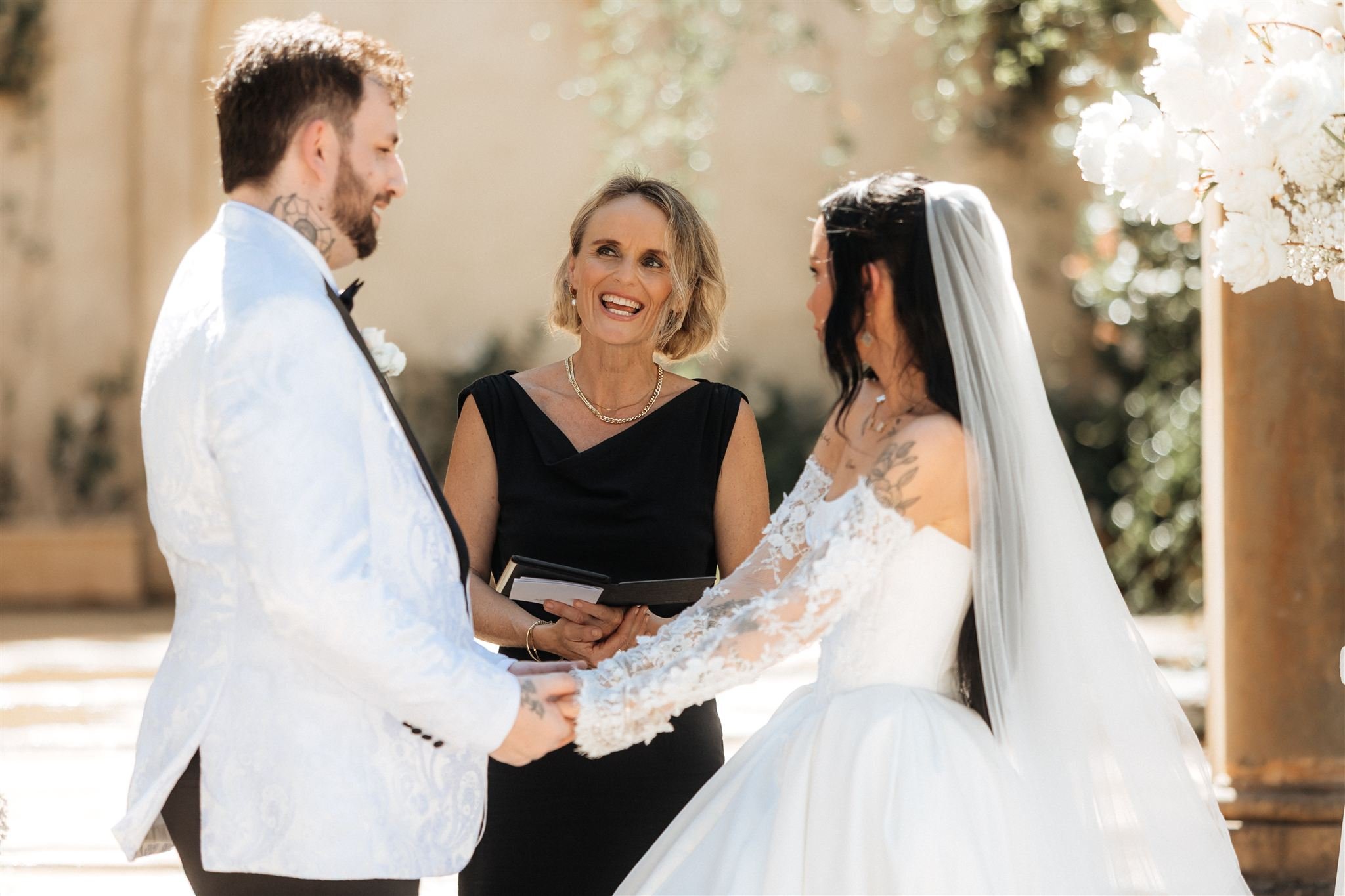 A couple getting married outdoors, with a woman officiating, holding hands and facing each other, during a wedding ceremony with white flowers and trees in the background.