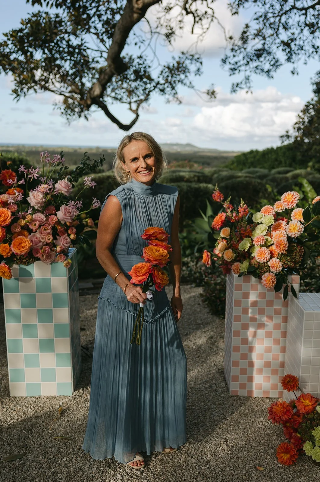 A smiling woman in a blue pleated dress holding a bouquet of orange flowers, standing among floral arrangements on checkered pedestals outdoors, with a large tree and a scenic landscape in the background.