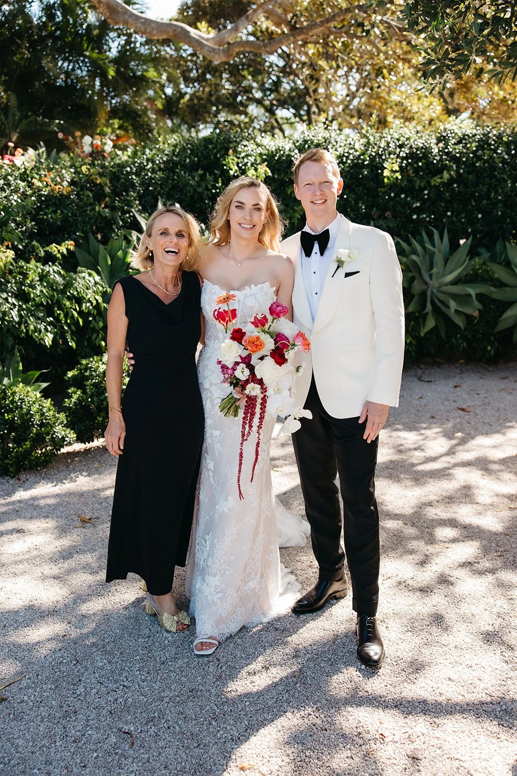 A wedding photo of three people: a woman in a black dress, a bride in a white wedding gown holding a bouquet of colorful flowers, and a groom in a white tuxedo jacket with a black bow tie, standing outdoors with greenery and trees in the background.