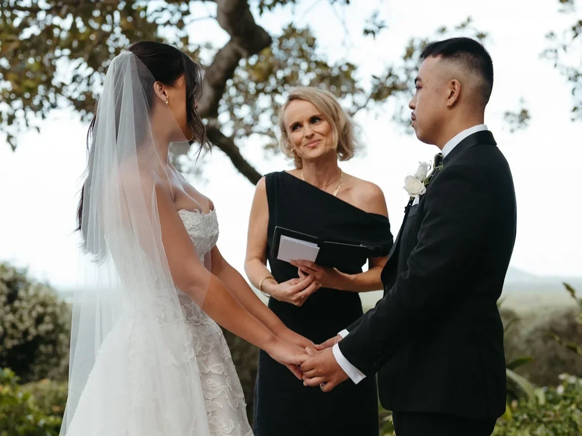 A wedding ceremony outdoors with a bride and groom holding hands, facing each other, with an officiant standing between them. The bride is wearing a white wedding dress with a veil, and the groom is dressed in a black tuxedo. The officiant, a woman in a black dress, holds a book and looks at the couple.