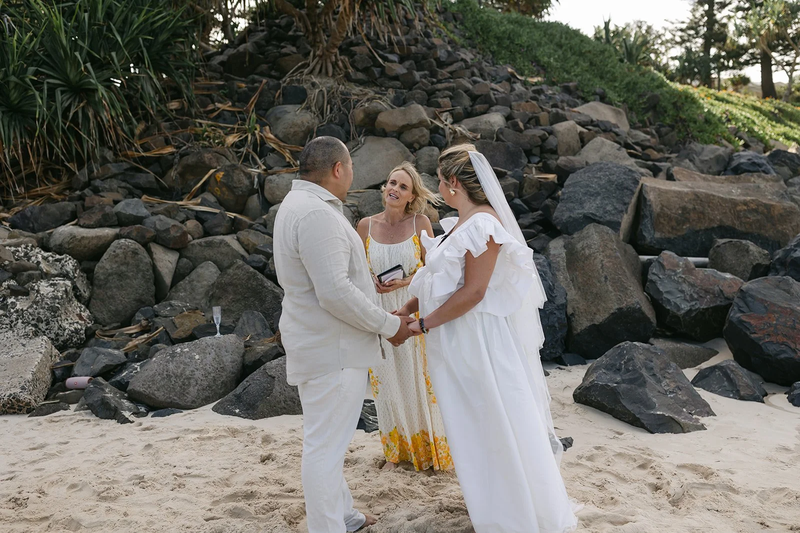 A couple getting married on a beach, holding hands and exchanging vows, with a woman officiating next to them and a rocky backdrop.