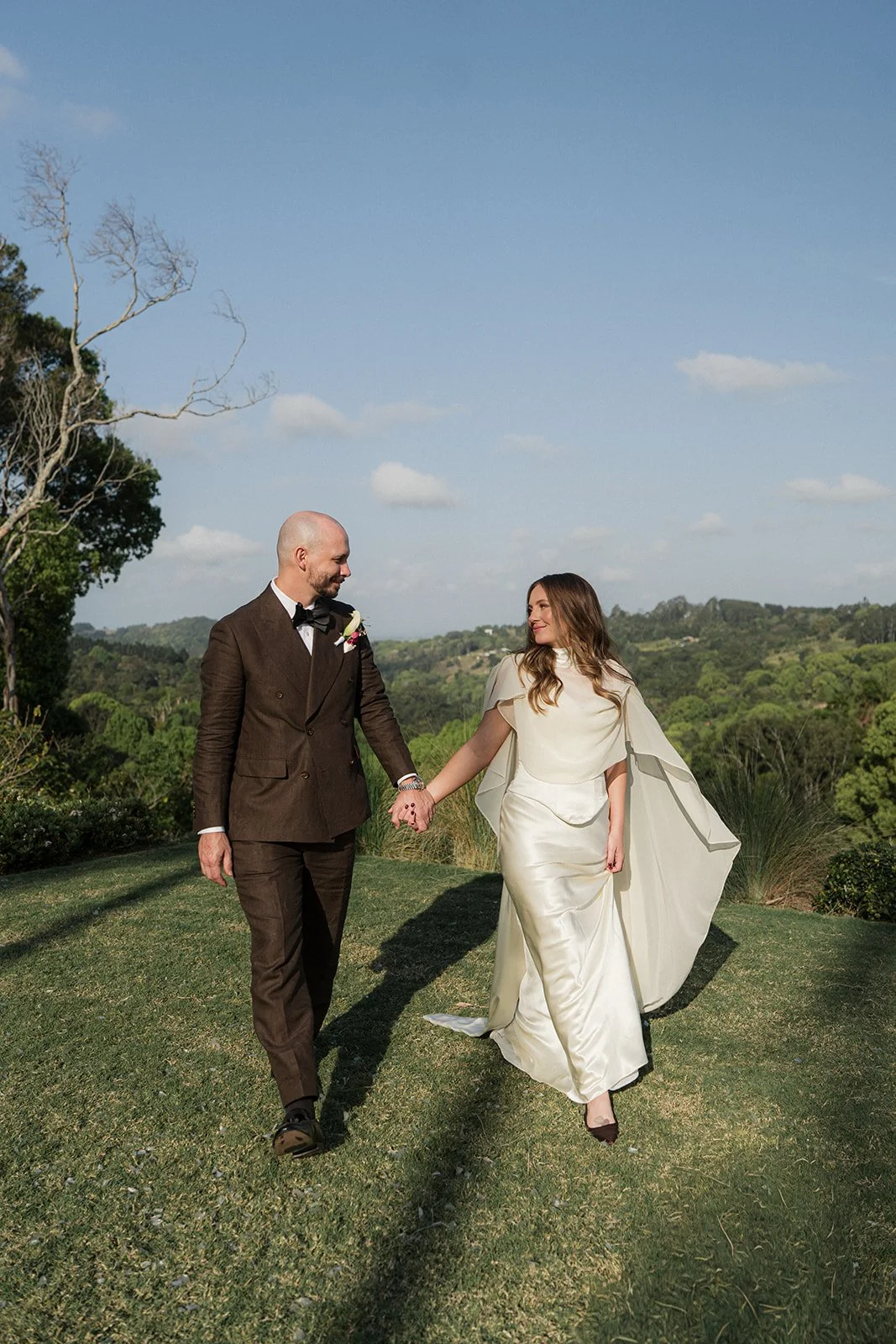 A bride and groom holding hands and walking outdoors on a grassy area with hills and trees in the background under a partly cloudy sky.