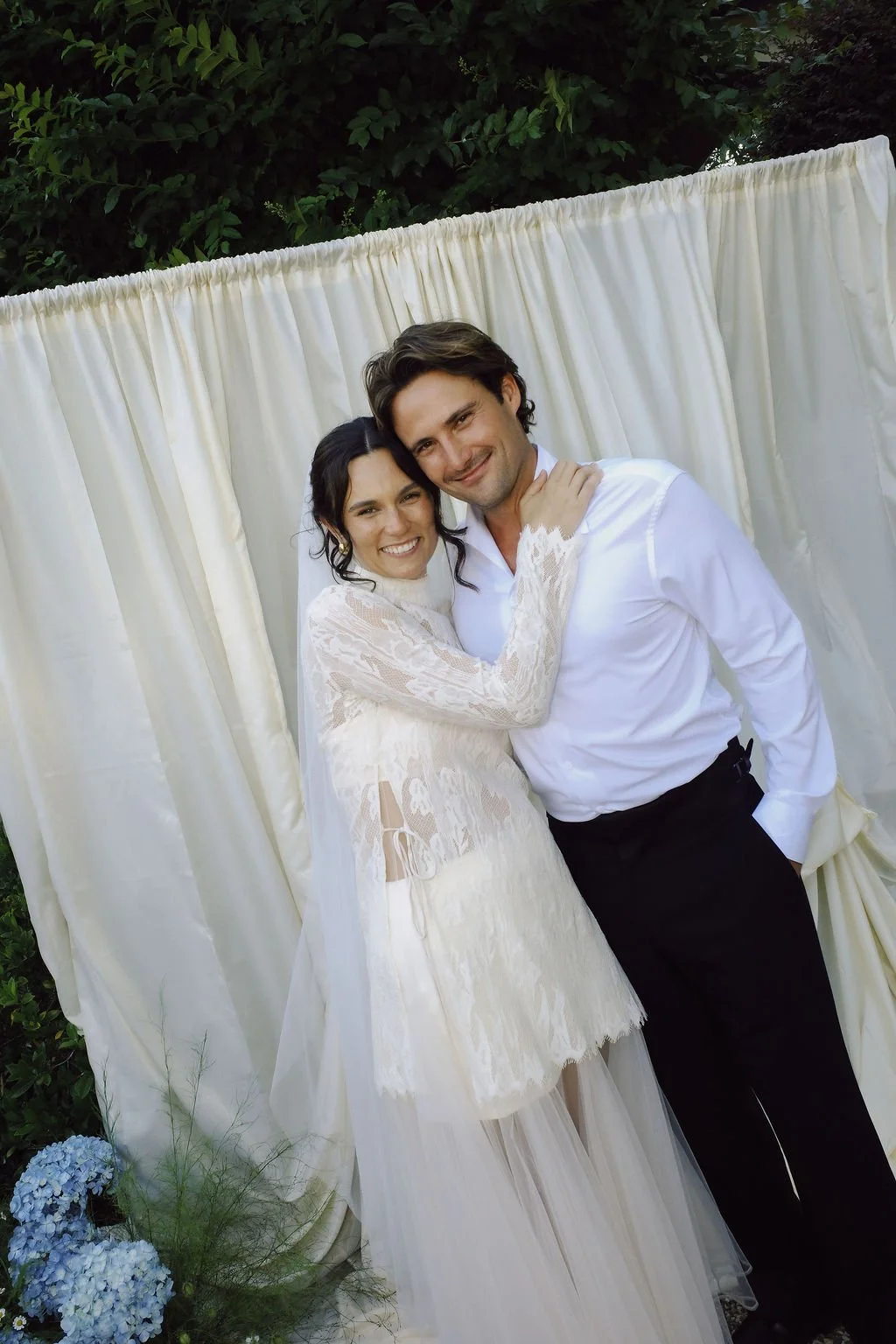 A bride and groom smiling and hugging each other at their wedding, posing in front of a cream-colored curtain with greenery behind and a small bouquet of blue hydrangeas on the ground.