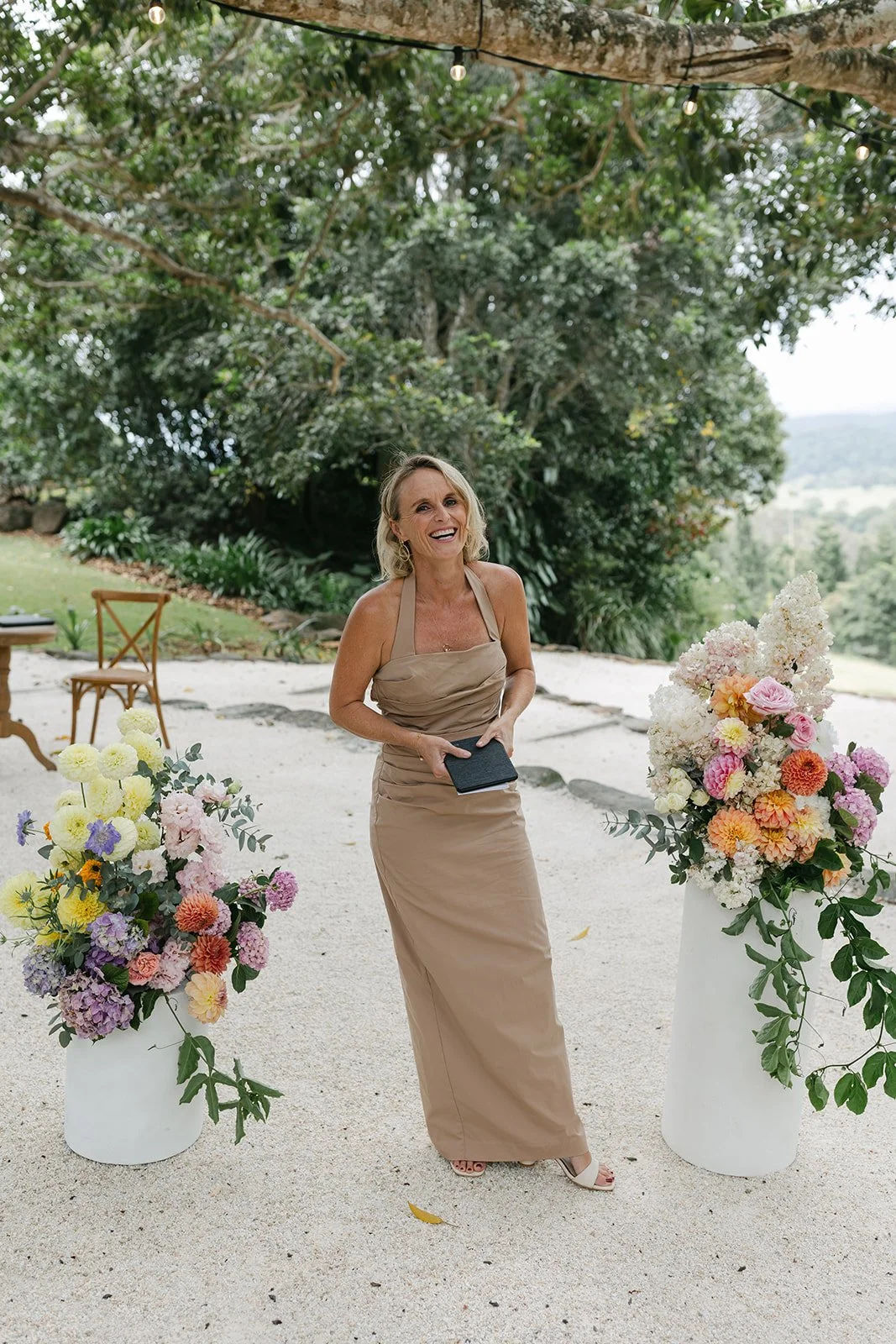 A woman in a beige dress is smiling at an outdoor event with large floral arrangements on white pedestals, surrounded by trees and greenery.