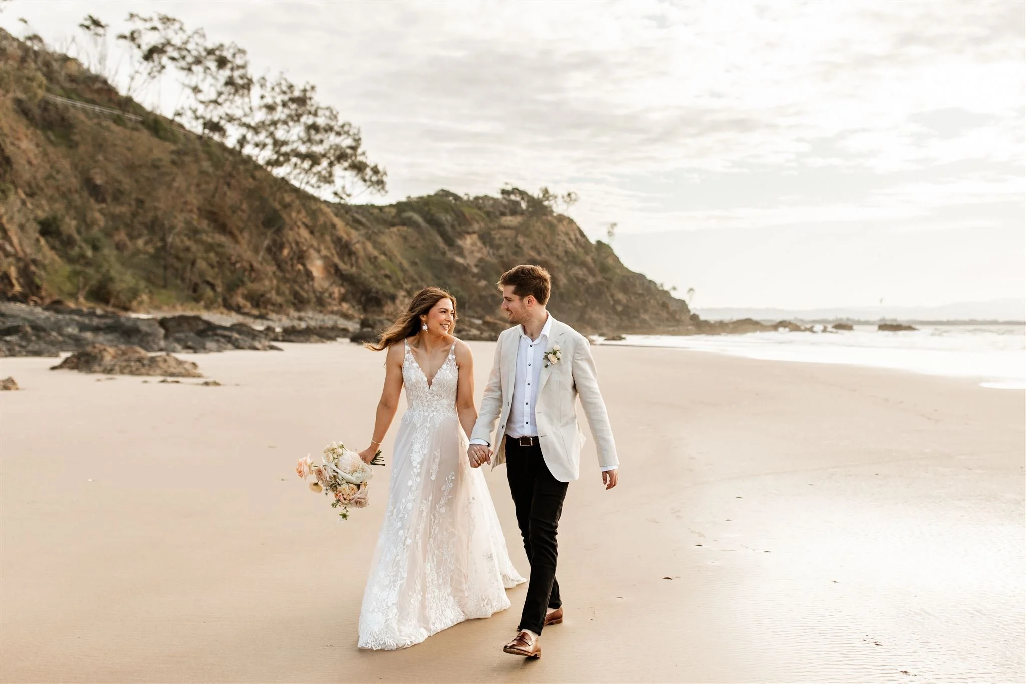 A bride and groom walking hand in hand on a beach, with cliffs and trees in the background.