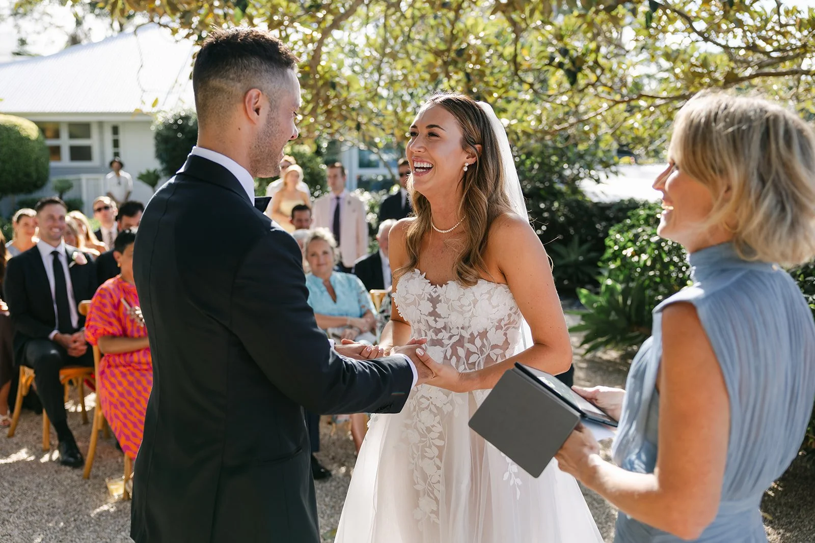 A bride and groom exchanging vows at an outdoor wedding ceremony, with guests seated in the background and trees providing shade.
