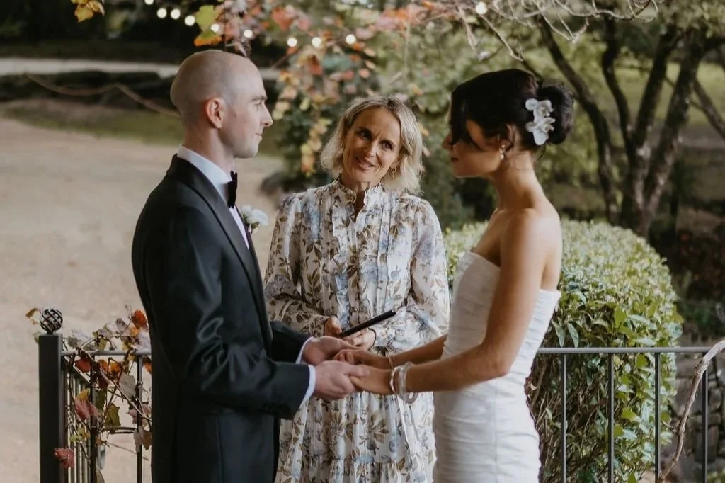 A couple getting married outdoors, holding hands and facing each other with an officiant standing behind them, in a lush garden setting.