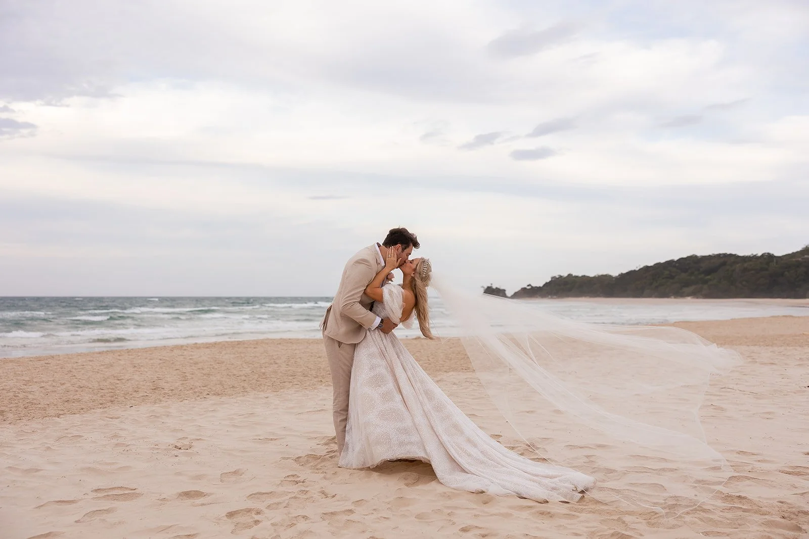 A bride and groom sharing a kiss on the beach, with the ocean and cloudy sky in the background.