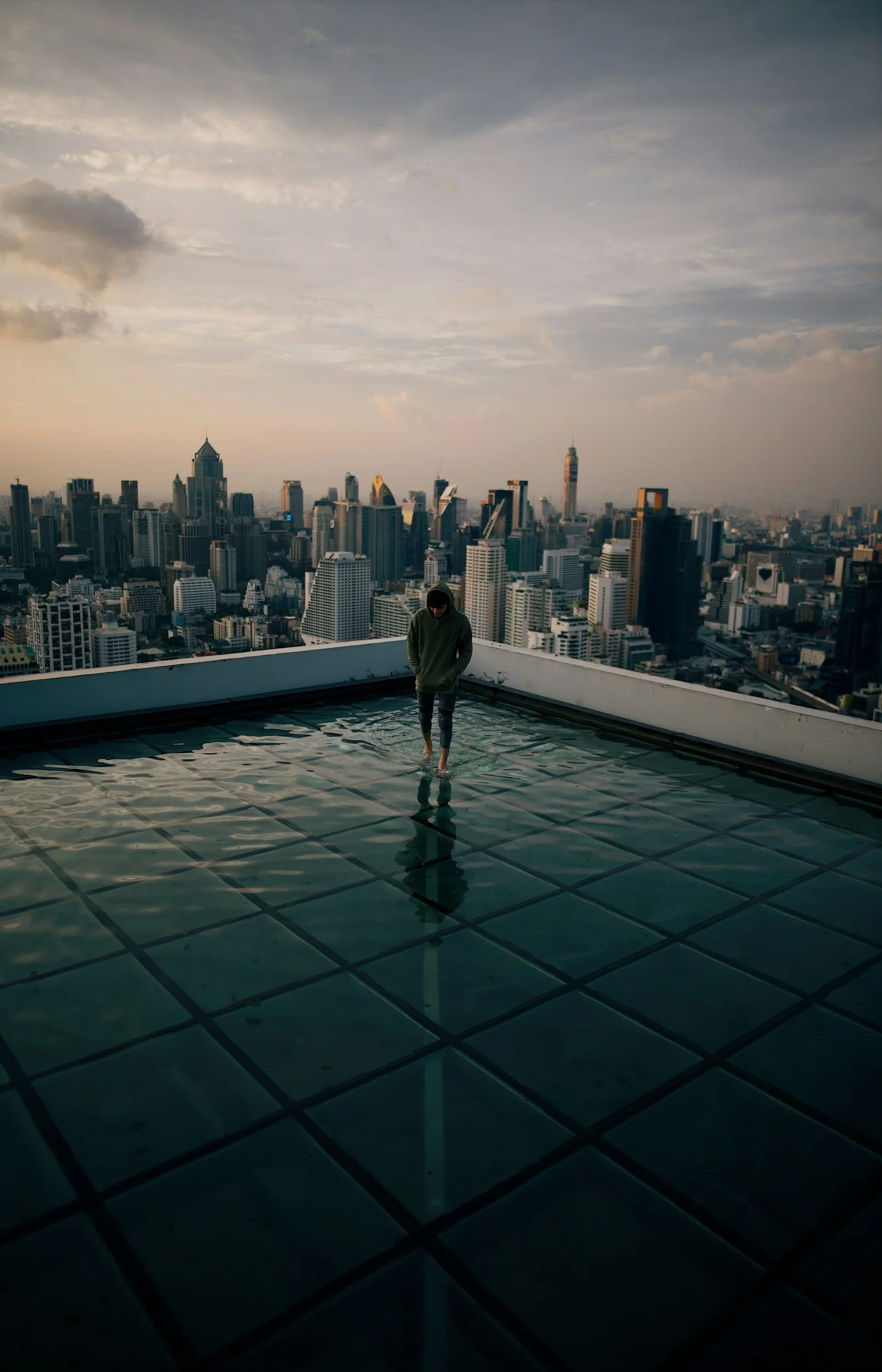 Person walking barefoot on a glass rooftop with a city skyline and cloudy sky in the background.