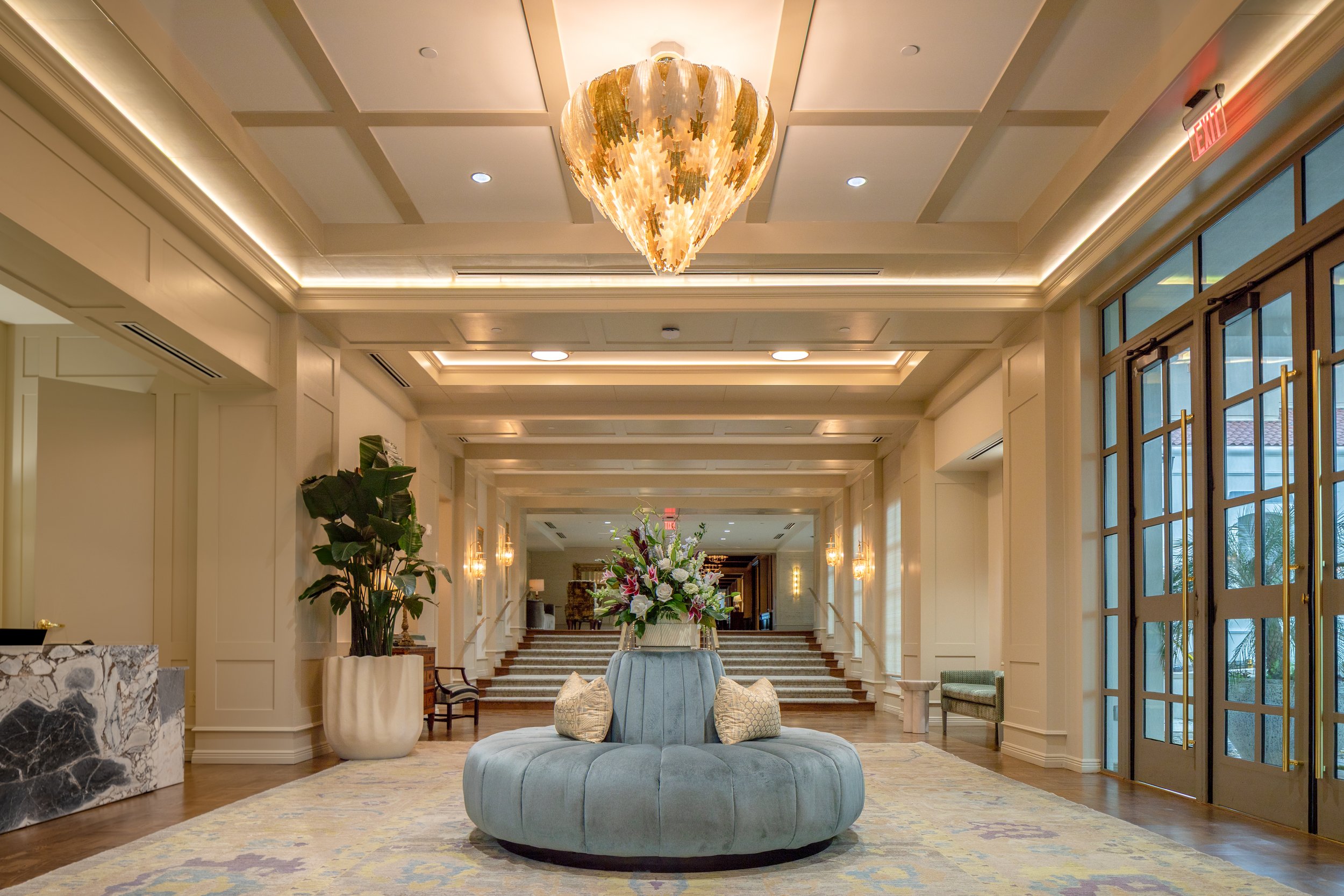Elegant hotel lobby with cream walls, a large floral arrangement on a circular velvet sofa, chandelier, potted plant, staircase, and glass entrance doors.
