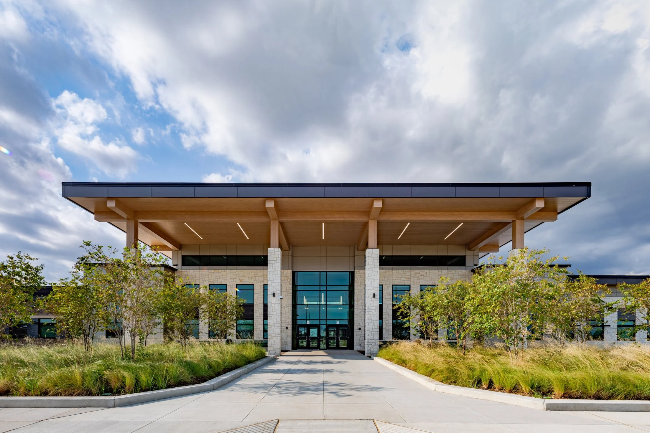 Modern building with a flat roof, glass entrance, and landscaped front walk and trees.
